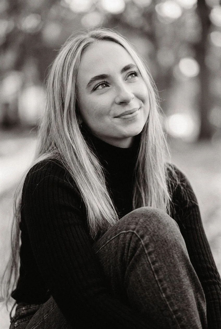 A young woman with long blonde hair sitting outdoors, looking up and smiling softly, with a blurred background of trees.