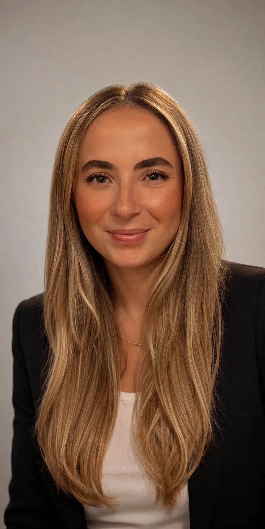 A young woman with long, wavy blonde hair and light complexion, smiling, wearing a black blazer and white top, against a plain grey background.