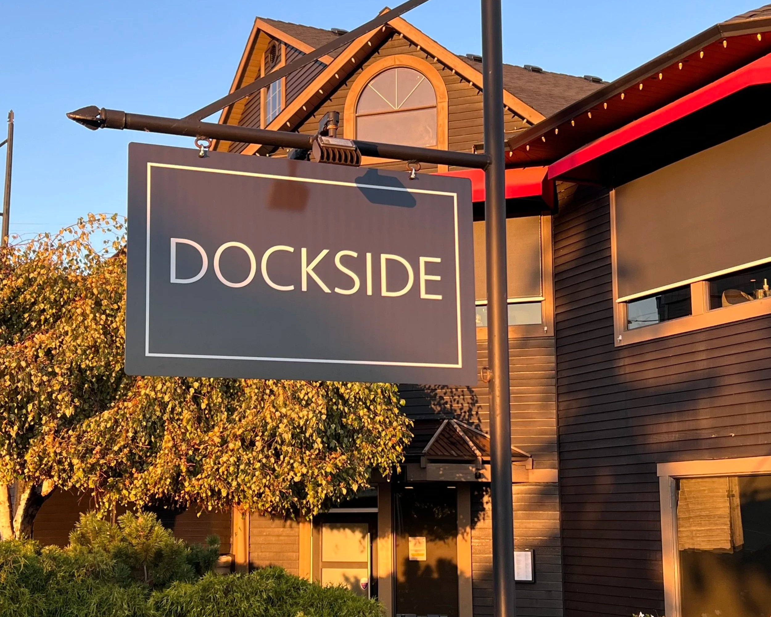 A sign that reads ‘Dockside’ hangs in front of a wooden building with dark brown siding, large windows, and a sloped roof under a clear blue sky at sunset.