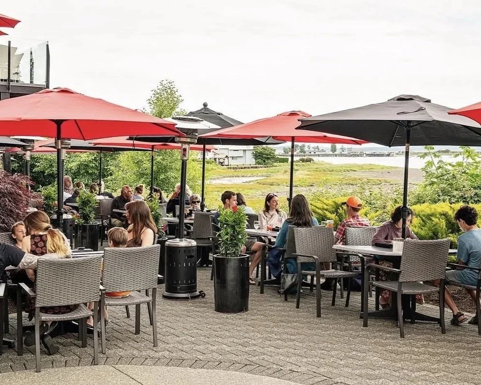 Outdoor restaurant patio with people dining under red and black umbrellas, surrounded by greenery, with a view of a river or lake in the background.