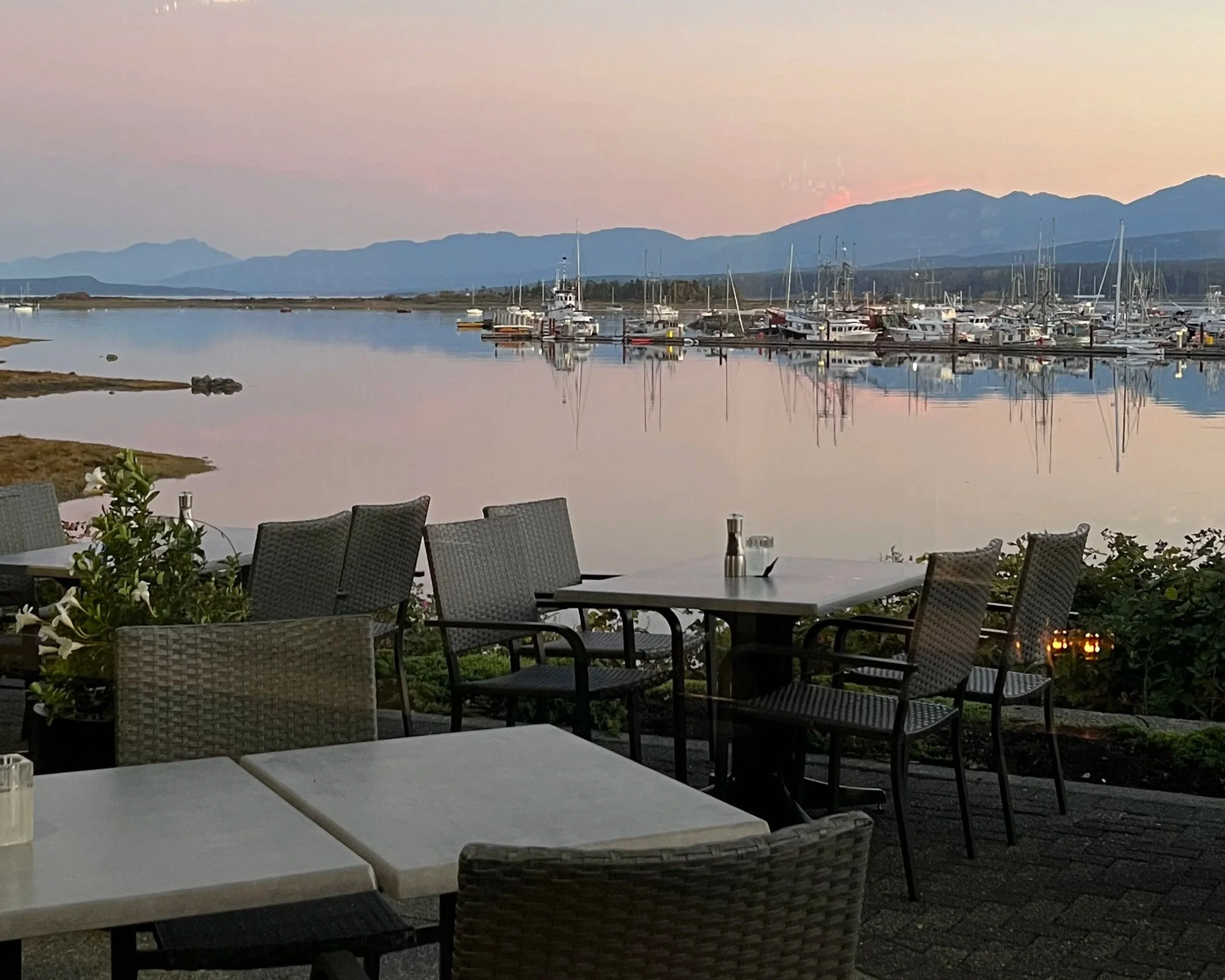 Outdoor dining area with tables and chairs overlooking a marina with boats and mountains in the background at sunset.