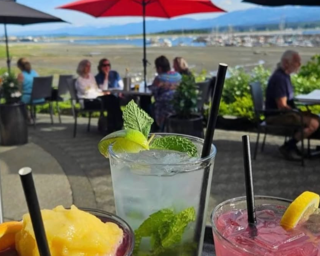 Three colorful cocktails with black straws on a table outdoors at a restaurant or café with red, black, and gray umbrellas, people sitting in the background, and a view of water and mountains in the distance.