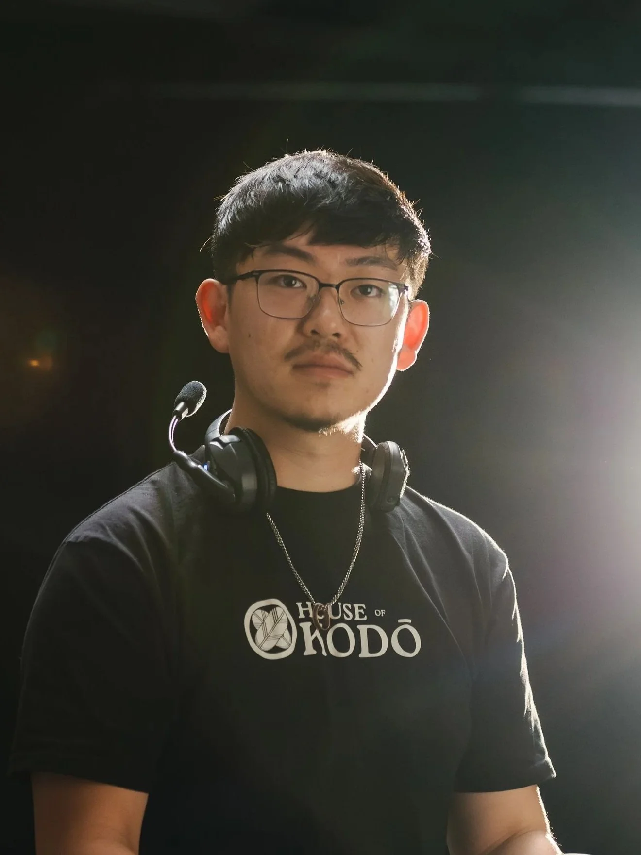 Young man with glasses, black t-shirt with 'HOUSE OF KODO' logo, headphones around neck, standing against dark background with bright light on right side.