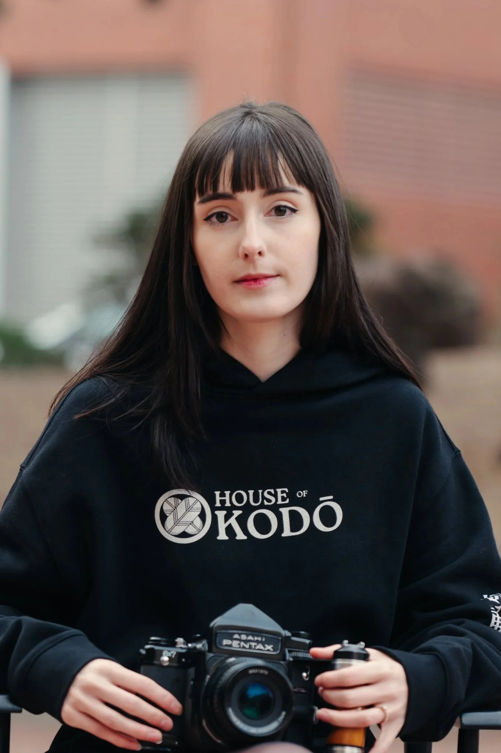 A young woman with long dark hair and bangs, wearing a black hoodie with 'House of Kodo' logo, holding a vintage film camera outdoors.
