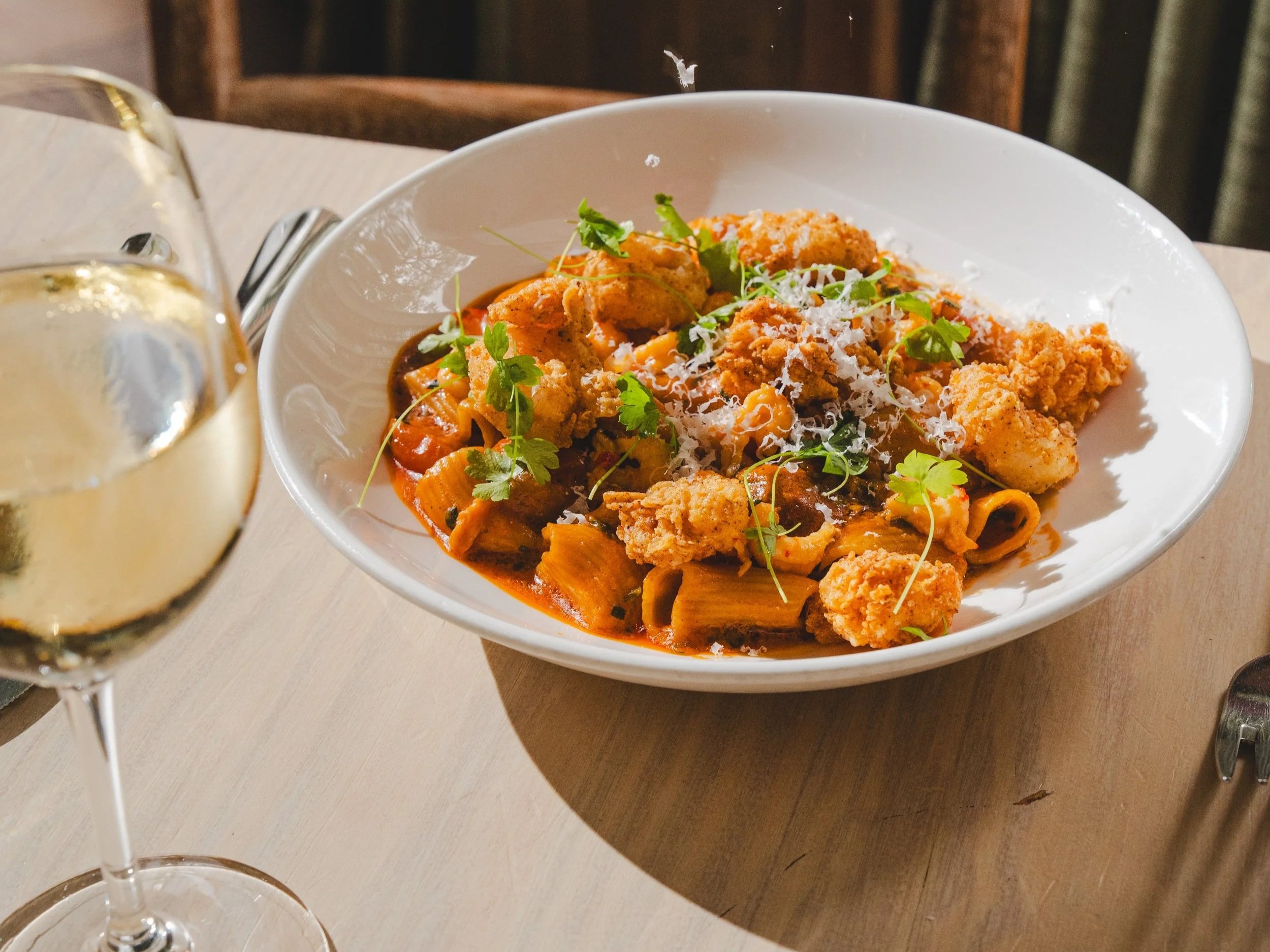 A glass of white wine and a bowl of Étouffée Pasta with Parmesan Cheese falling on top. 