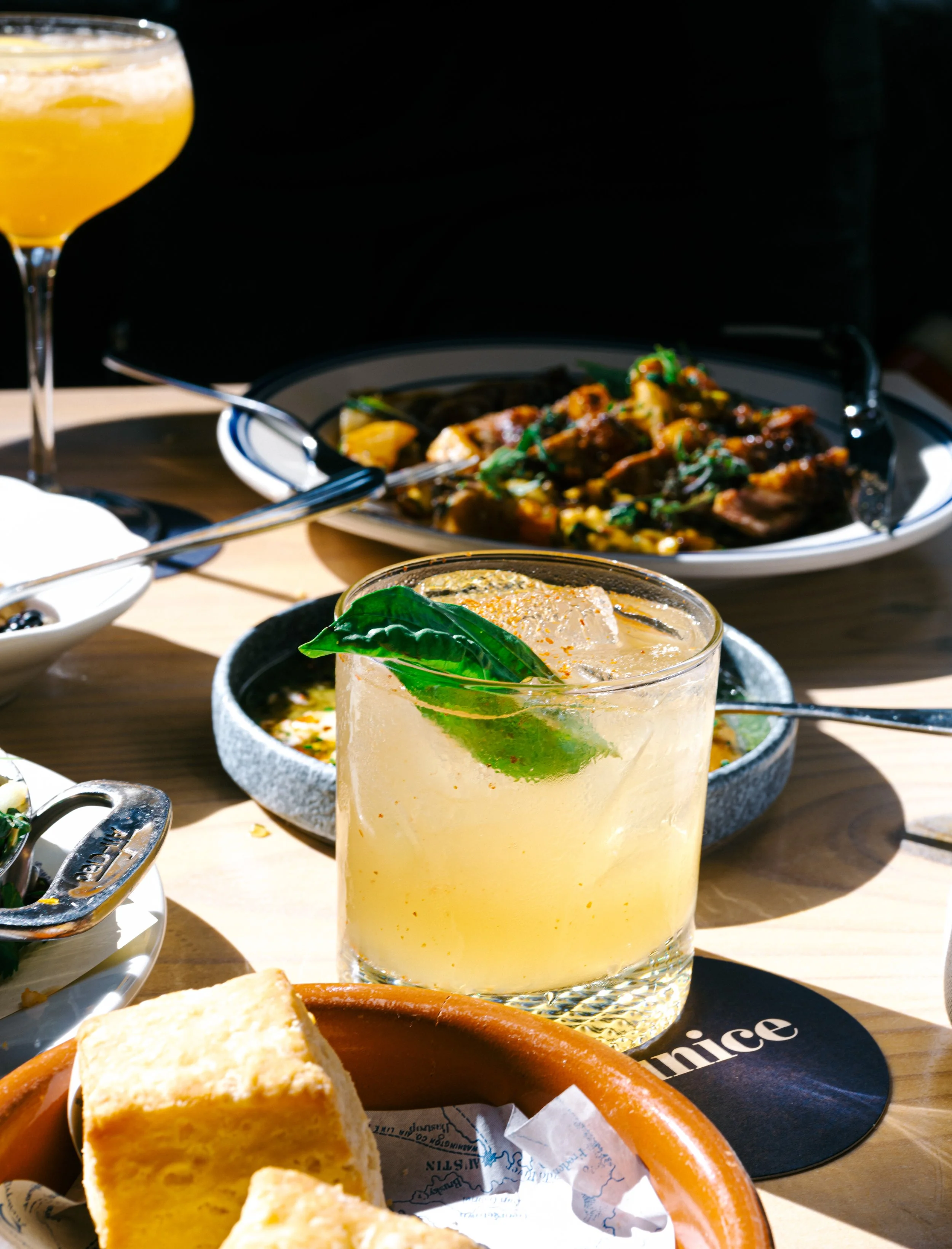 Close-up of a cocktail garnished with basil leaf, with dishes of food in the background on a wooden table.
