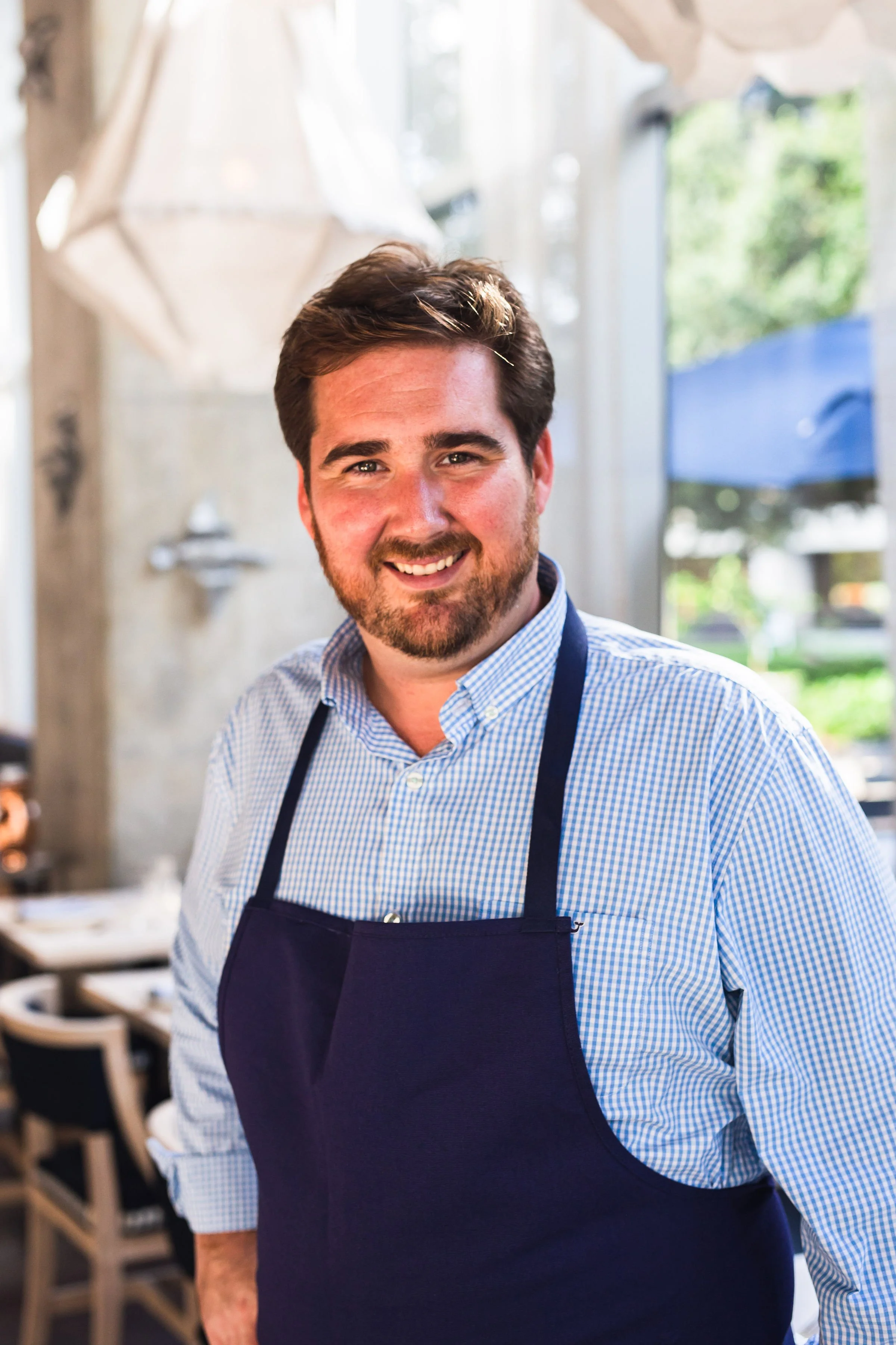A smiling man with brown hair and a beard, wearing a blue and white checkered shirt and a dark apron, standing inside a bright, airy restaurant or cafe with tables and chairs in the background.