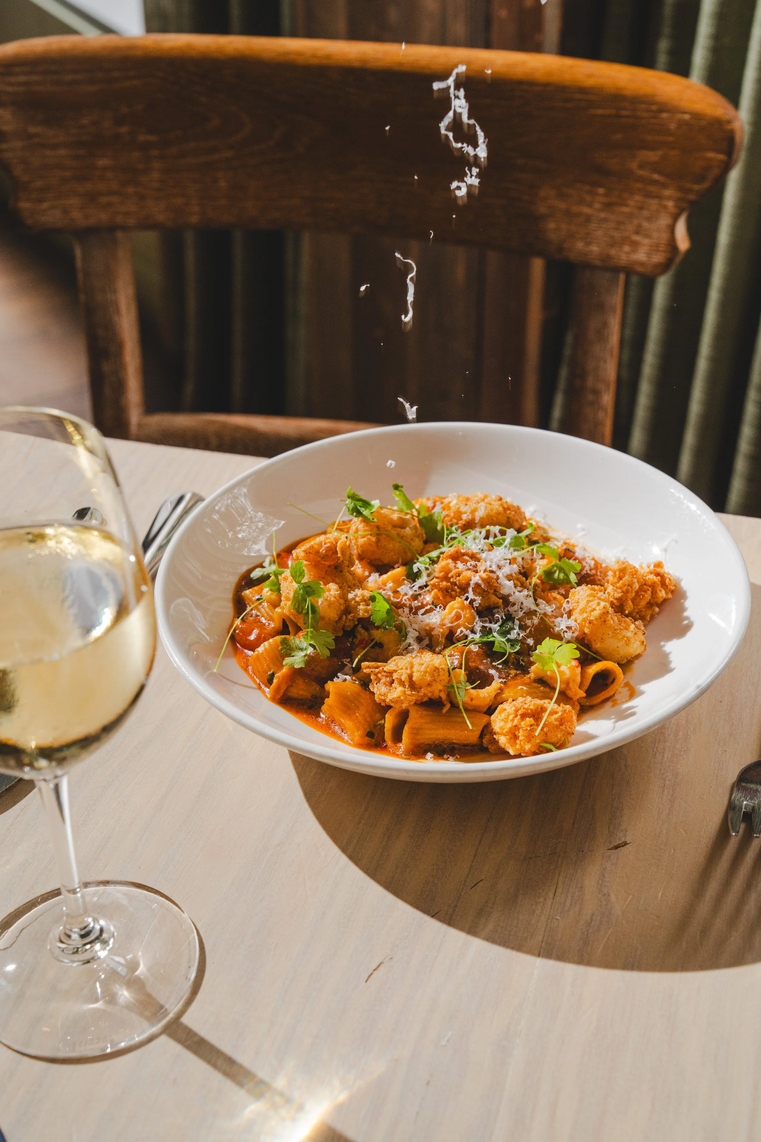 A glass of white wine and a bowl of Étouffée Pasta with Parmesan Cheese falling on top. 