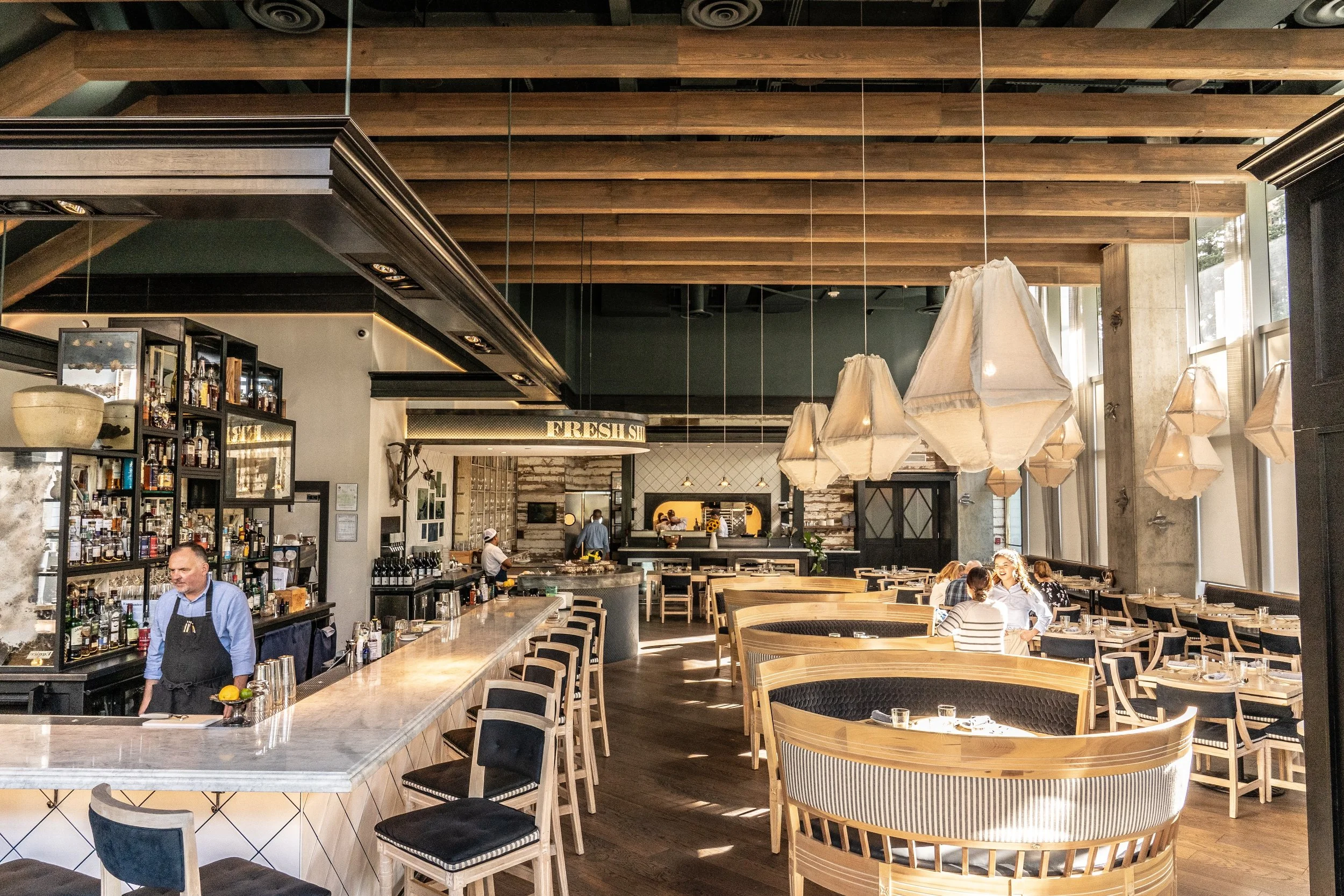 View of the main dining room looking towards kitchen from the main entrance. There is a bar with a bartender to the left, two women talking to the right. Further into the dining room there is a Raw Bar on the left with a staff member working.