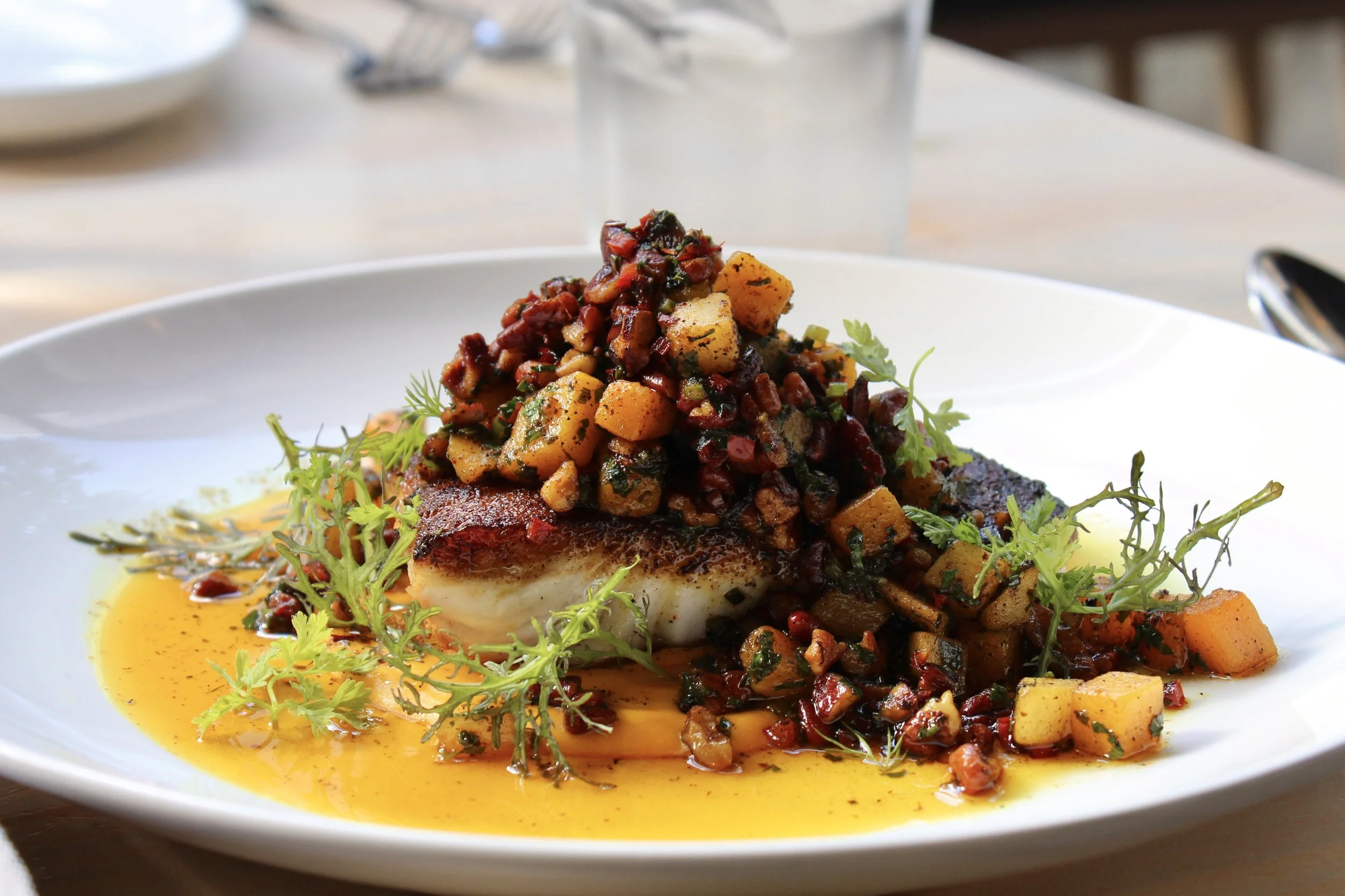 A plated dish consisting of a piece of white fish topped with a colorful mixture of squash, diced vegetables, and herbs, garnished with fresh greens, and surrounded by a yellow sauce.