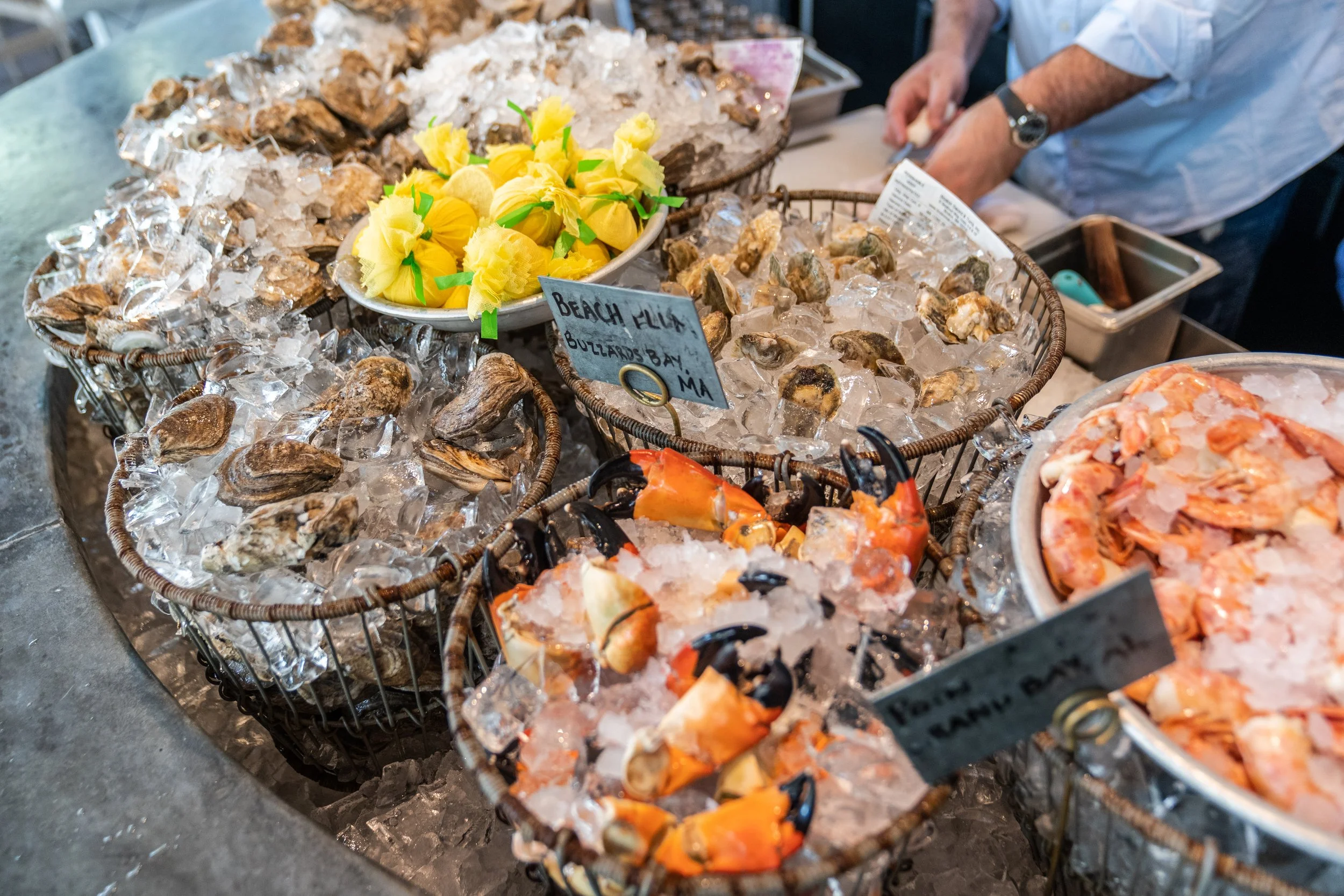 Raw oysters, crab claws, and shrimp chilled on ice at at Raw bar. In the background, an oyster is being shucked. 