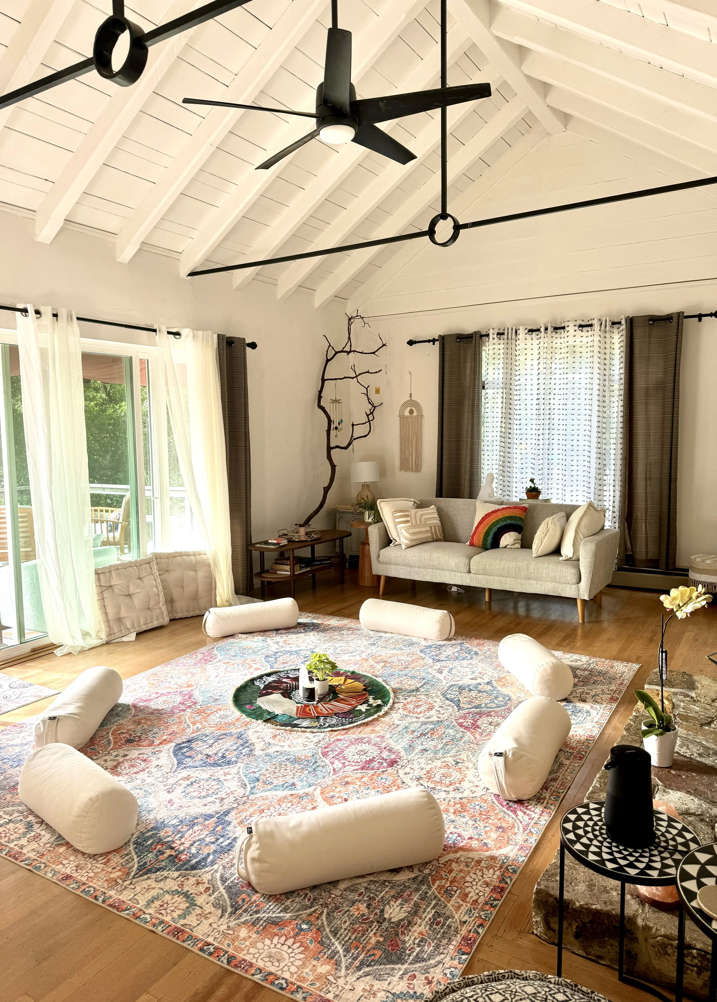 Cozy living space with vaulted white ceiling, modern ceiling fan, patterned area rug, circular arrangement of floor cushions, a light grey sofa with colorful pillows, large windows with sheer curtains, a decorative branch on the wall, and a small side table with plants.