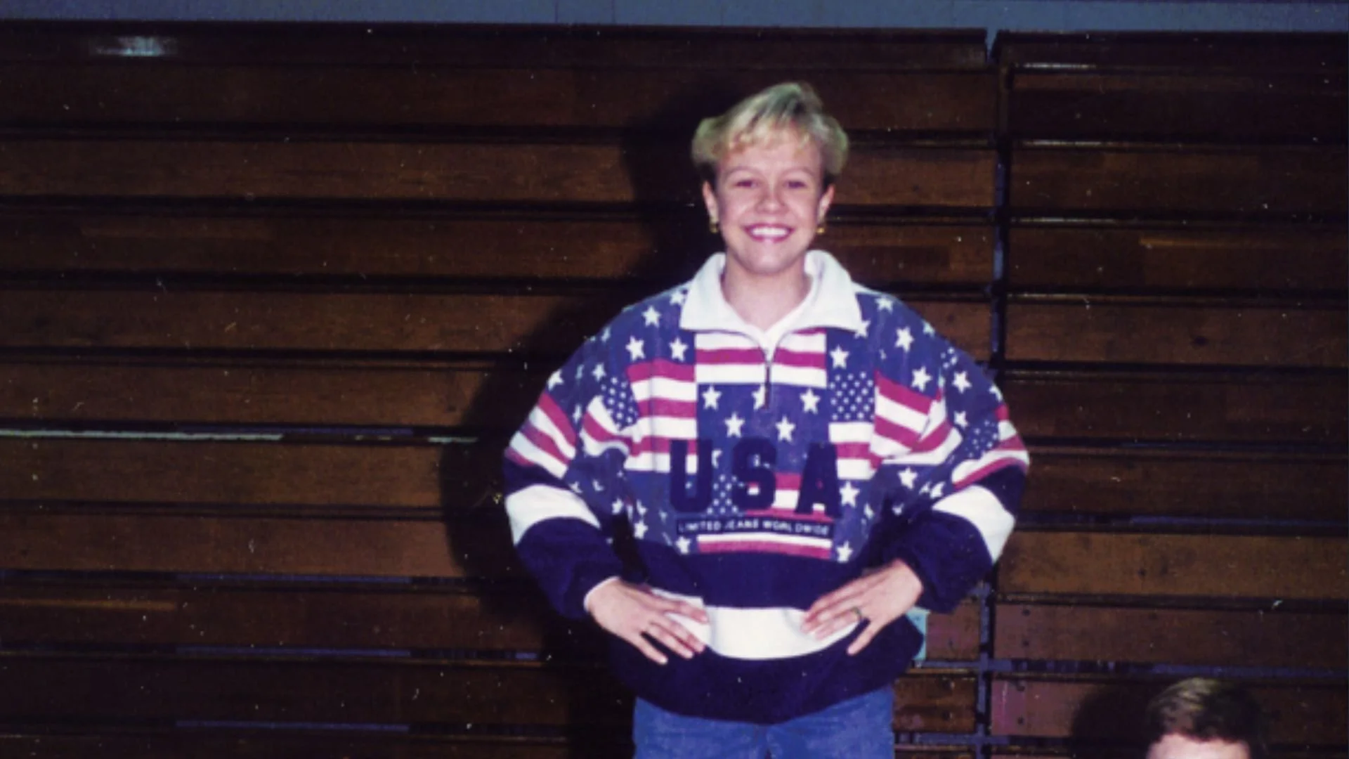 A woman standing in front of a wooden backdrop, wearing a U.S.A. themed sweatshirt with stars and stripes, smiling with her hands on her hips.