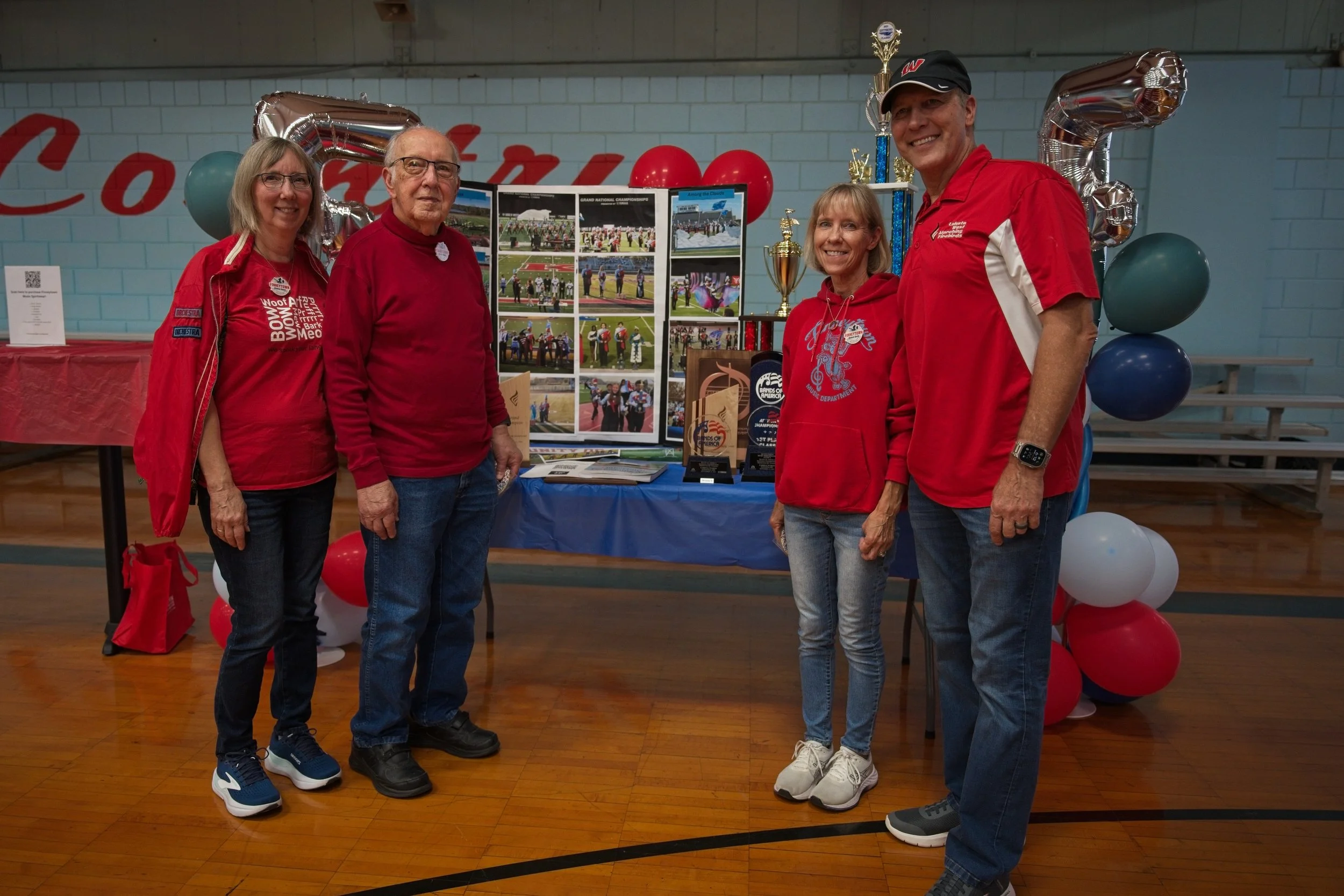 Five people standing in a gymnasium in front of a display table with photos, trophies, and medals. The background has balloons and a lettered banner. The people are wearing red and casual clothing.
