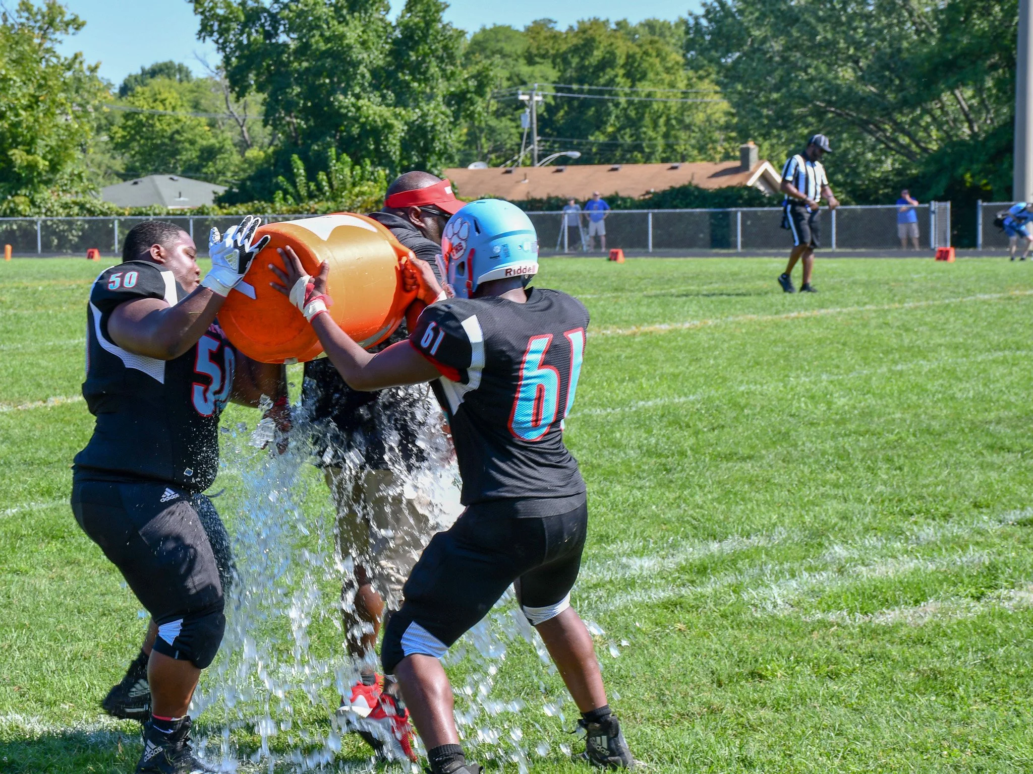 Football players practicing with a tackling dummy during a game, with other players and coaches in the background on a sunny day.