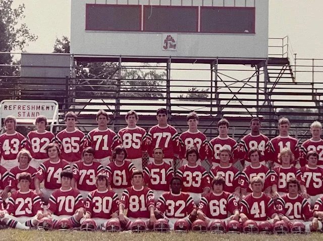 A youth football team in red and white uniforms posing for a team photo on a football field with bleachers in the background.