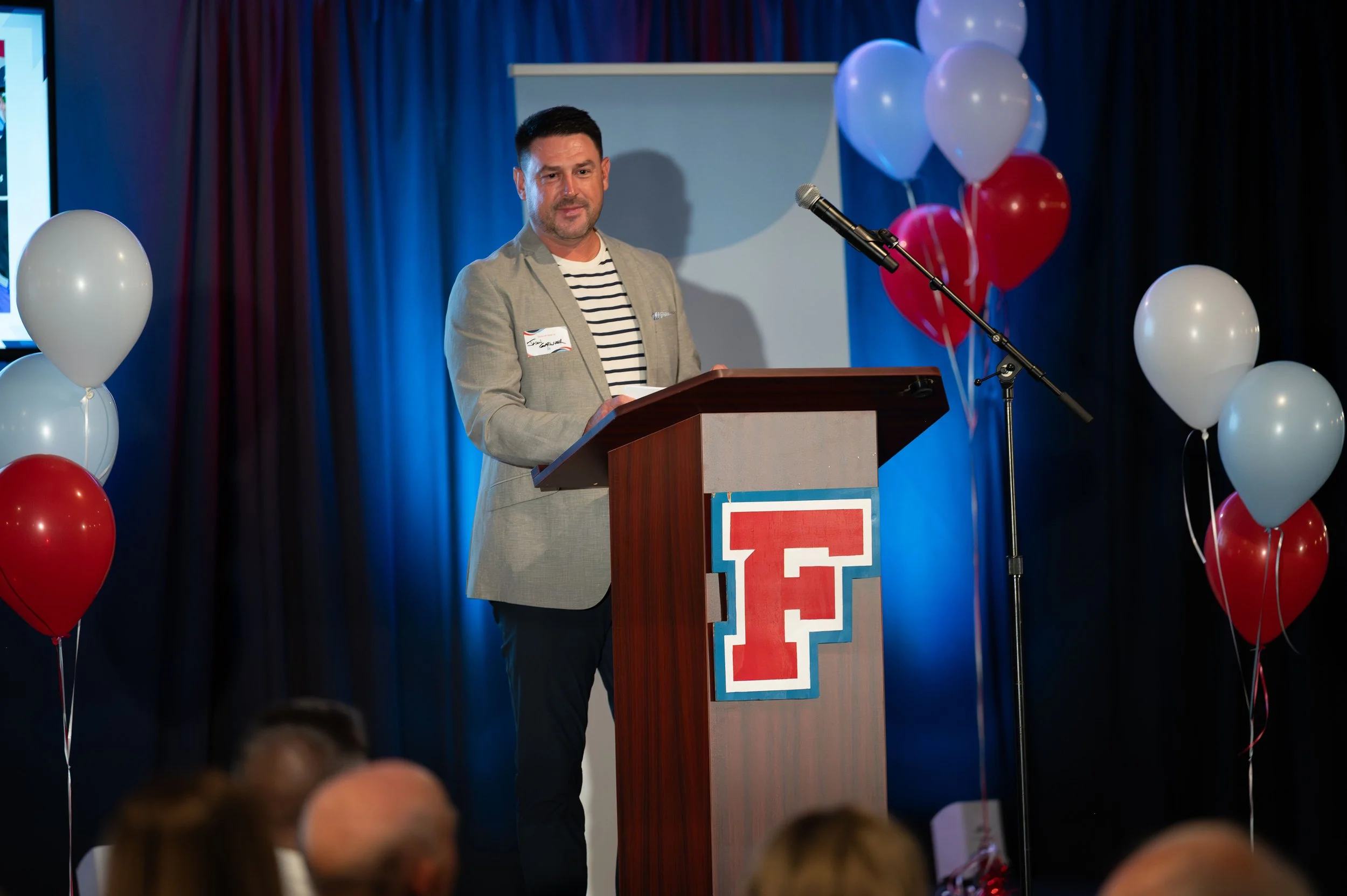 A man is standing at a podium during a speech or presentation at an event, with balloons and a large letter 'F' on the front of the podium.