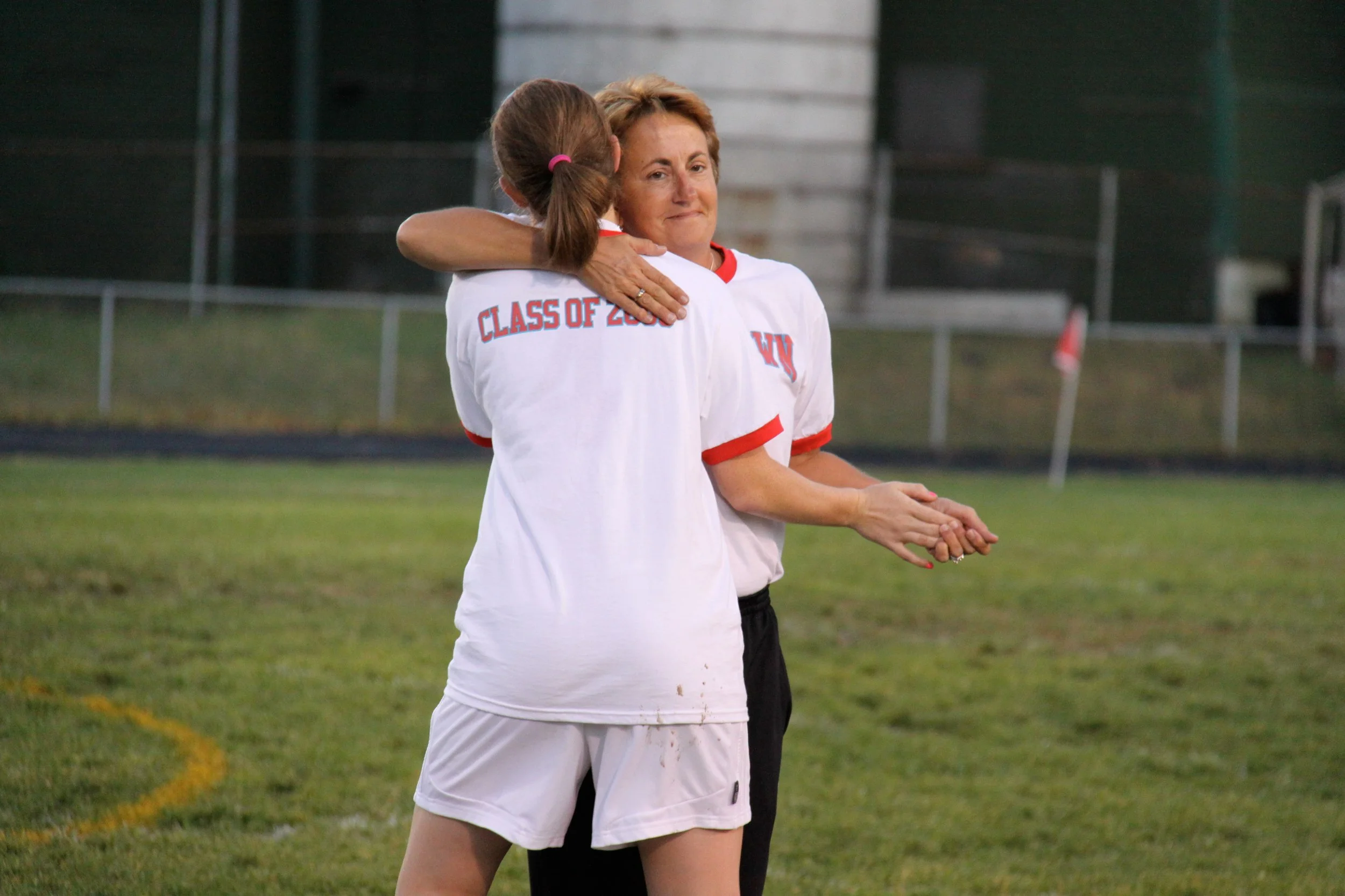 A young female athlete hugging an older woman on a sports field, both wearing white sports jerseys with red accents. The older woman appears emotional, and the young athlete has her back to the camera. The setting is a grassy field with a fence and a football corner flag in the background.