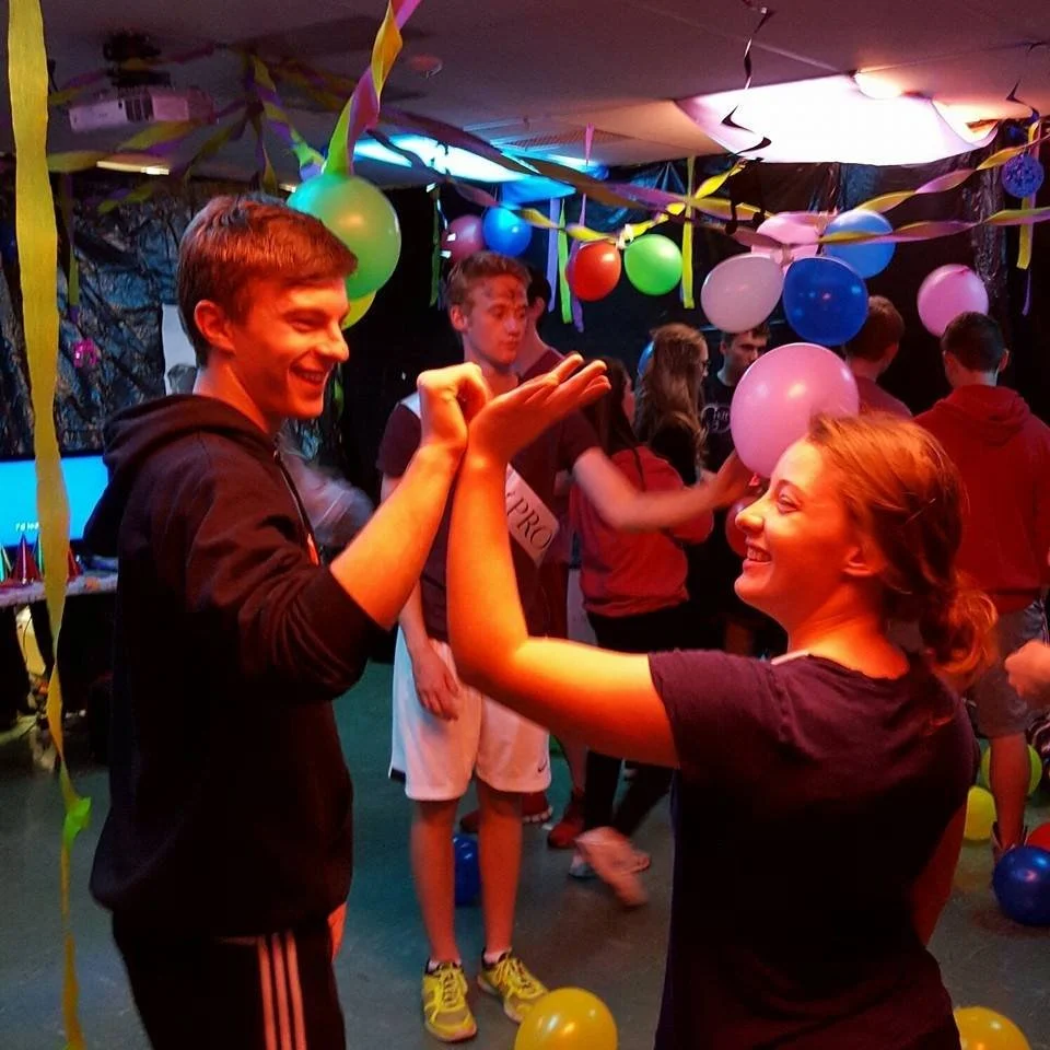 A group of teenagers dancing and celebrating at a party decorated with balloons and streamers.