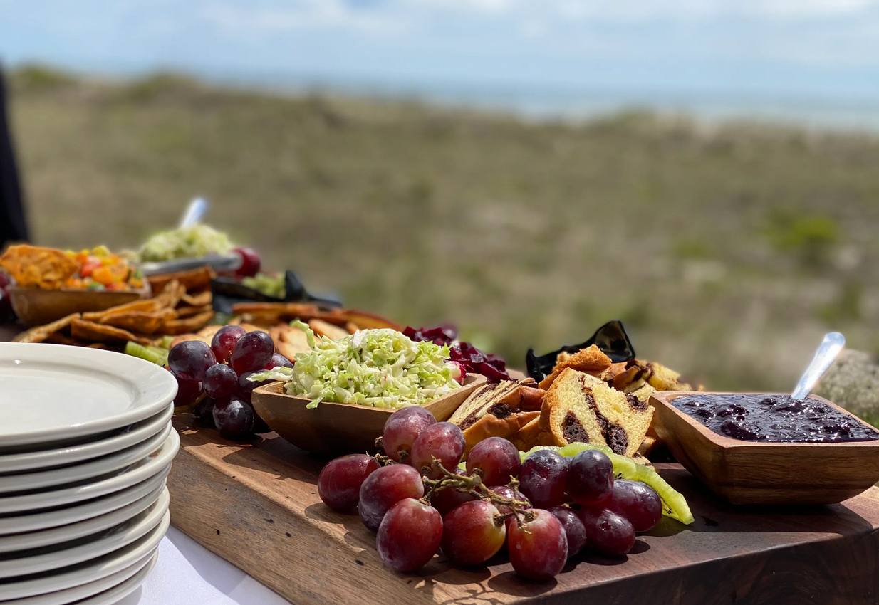 Assorted desserts and fresh grapes on a wooden table outdoors, with blurred landscape in the background.