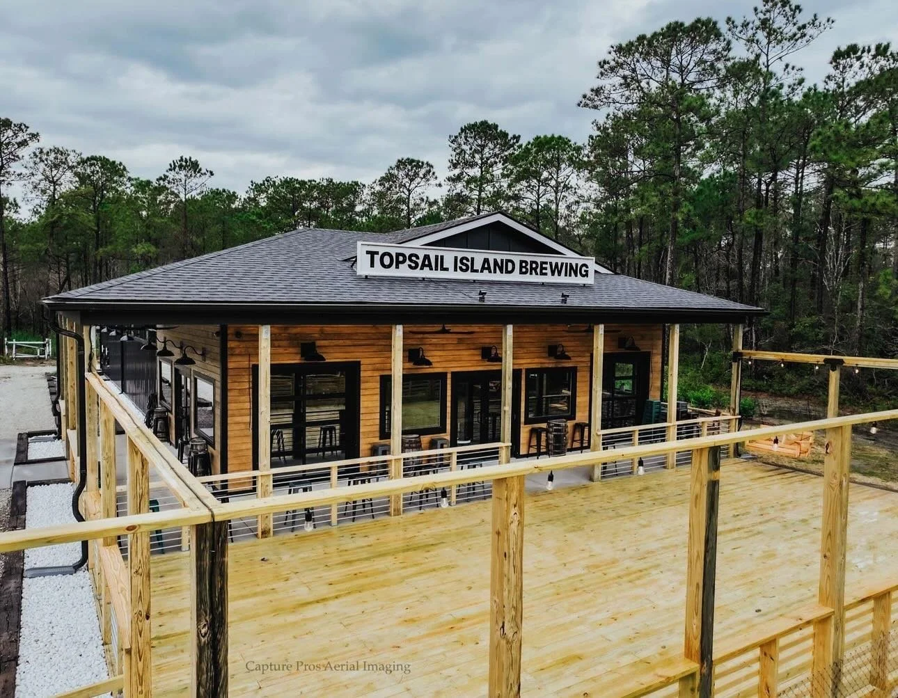 A building with a sign reading 'Topsail Island Brewing,' with a spacious wooden deck in front, surrounded by trees and cloudy sky.