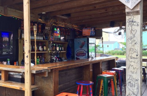 Empty bar with colorful stools, shelves with bottles, and a large window showing outdoor scenery.