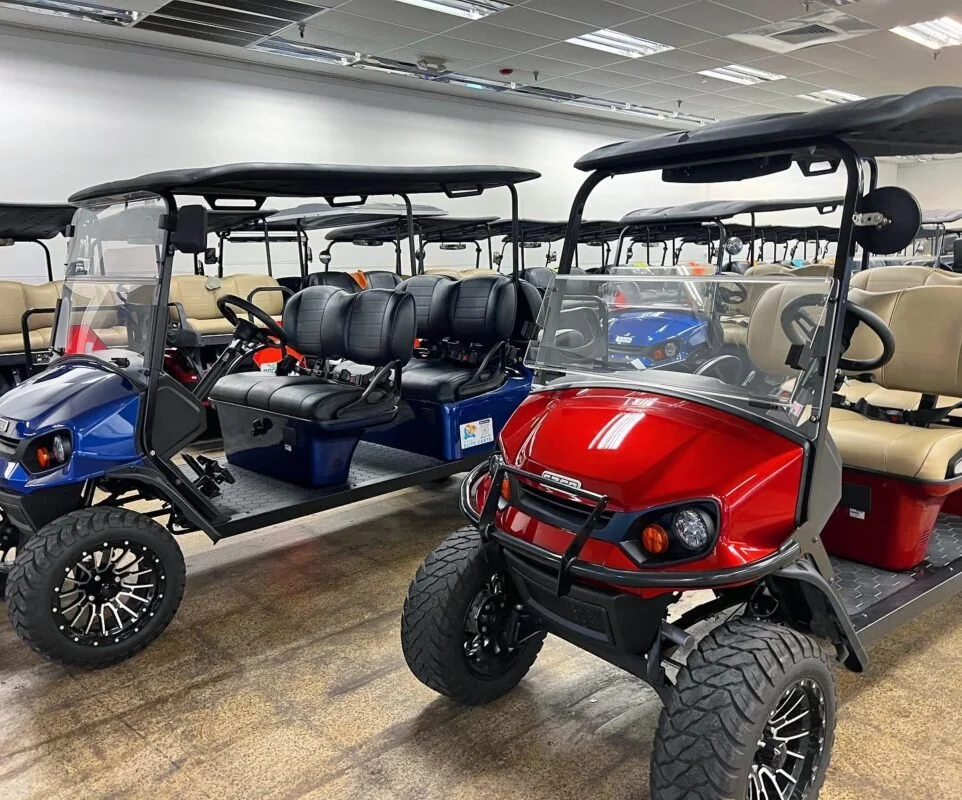 Multiple golf carts parked indoors; front one is red with large tires, the others are blue and beige, all with roofs and windshields.