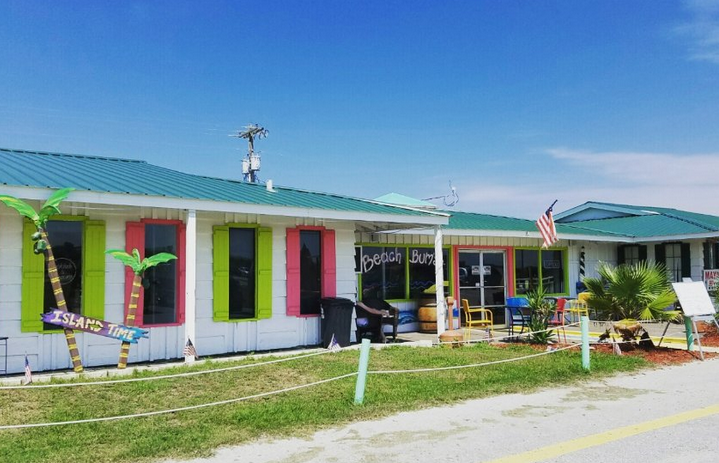 Colorful beach-themed building with green and pink window shutters, inflatable palm trees, and outdoor seating area, with a blue sky and American flag.