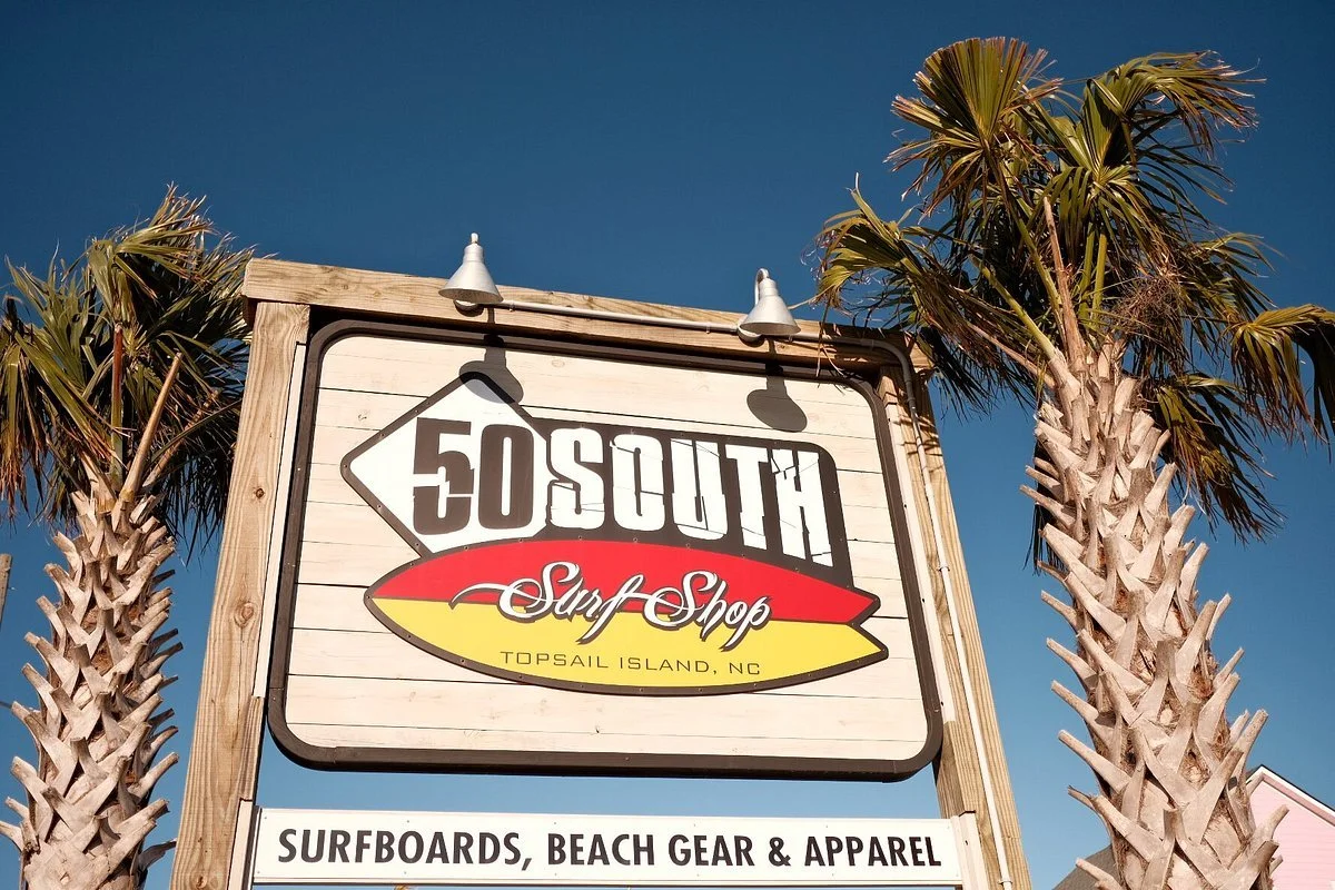 Sign for 50 South Surf Shop with palm trees on either side, indicating it sells surfboards, beach gear, and apparel in Topsail Island, North Carolina.