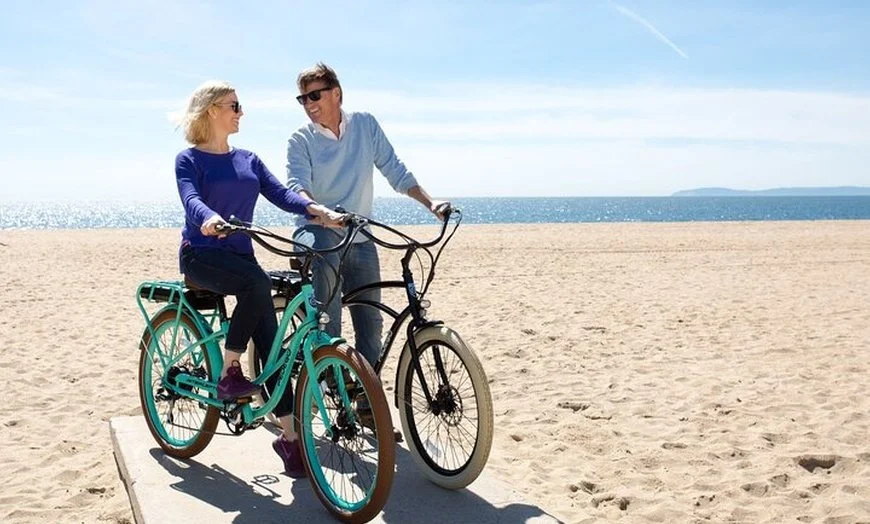 A woman sitting on a teal and black tandem bicycle at the beach, with a man standing beside her, both smiling and holding the bike, under a blue sky with calm ocean in the background.