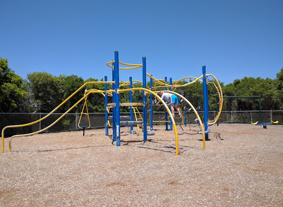 Empty playground with blue and yellow climbing structures and a gravel surface, surrounded by trees and a clear blue sky.