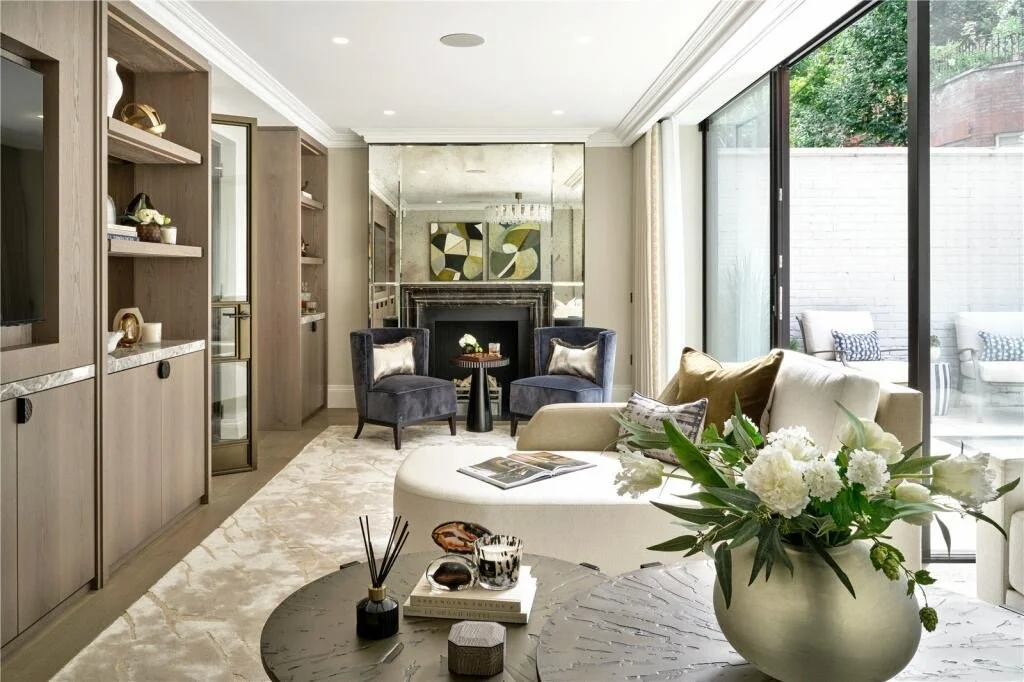 Bright living room with large sliding glass doors opening to outdoor patio, beige sofa with pillows, two dark blue armchairs near fireplace, built-in shelves, coffee table with books and decor, and floral arrangement.