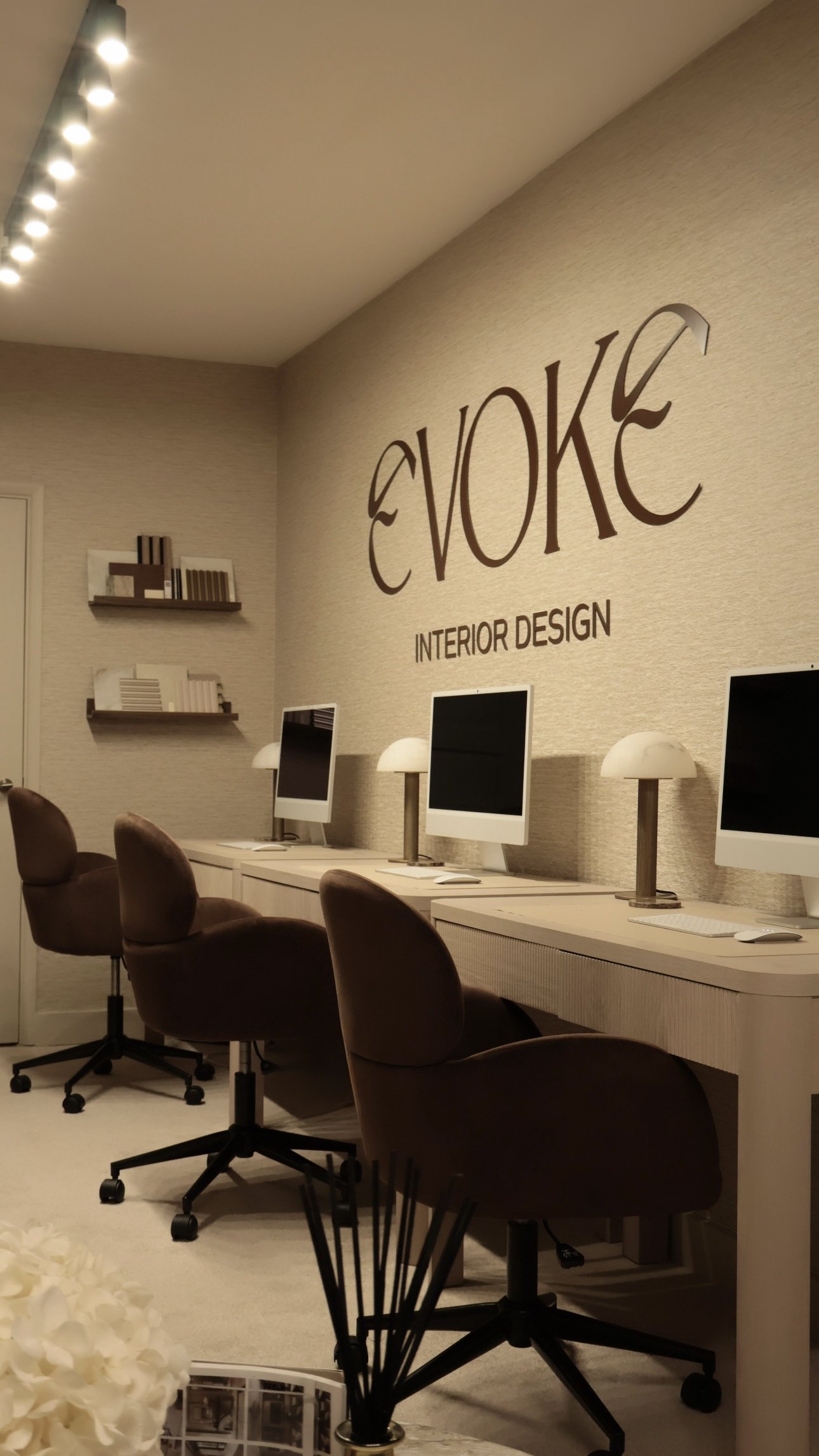 Interior design office with three computer stations, brown chairs, wall-mounted shelves with decor, and the logo 'EVOKE Interior Design' on the wall.