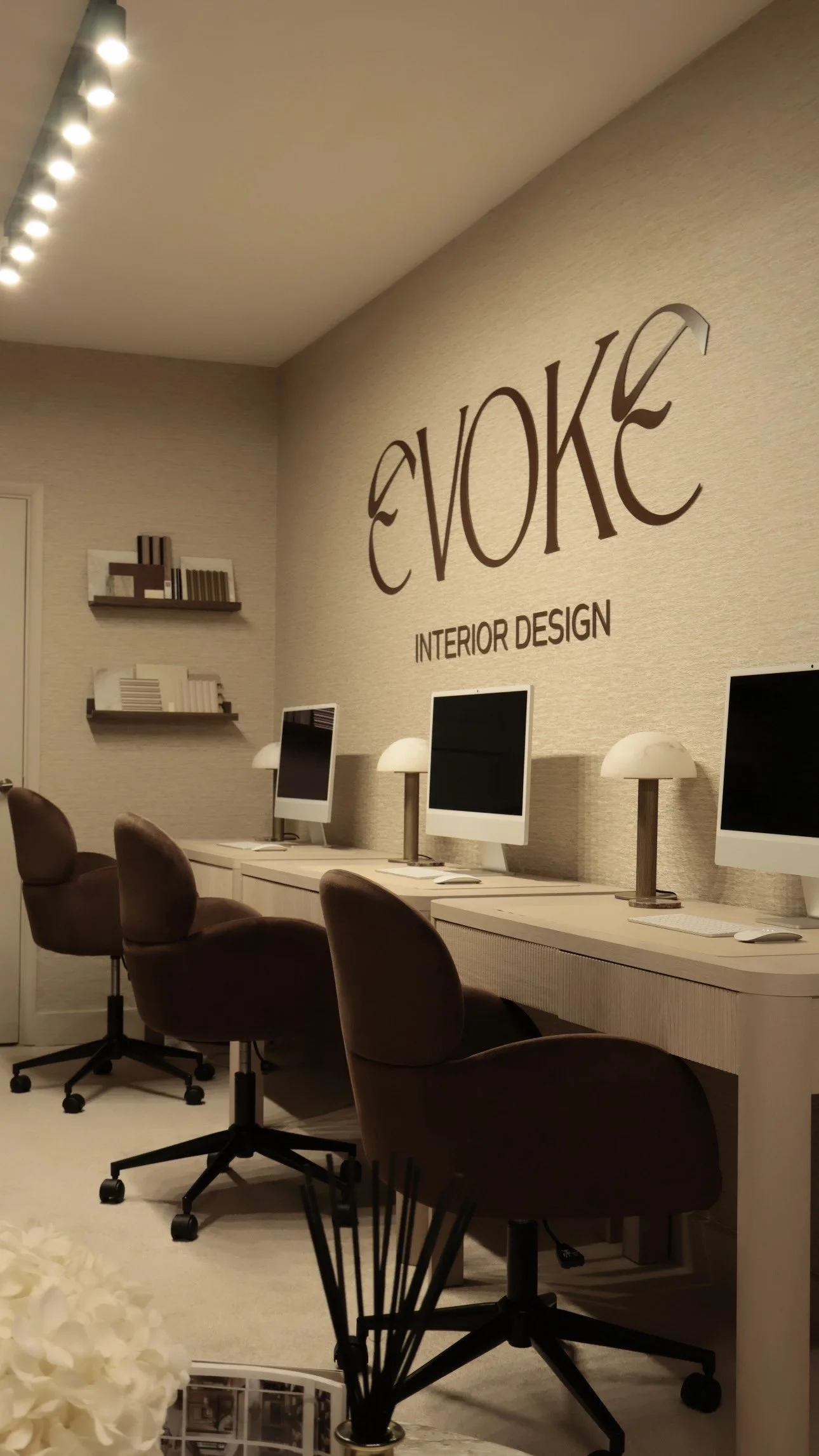 Interior of a modern office with four Apple iMac computers on a white desk, brown office chairs, wall-mounted lamps, and a logo on the wall that reads 'Evoke Interior Design'.