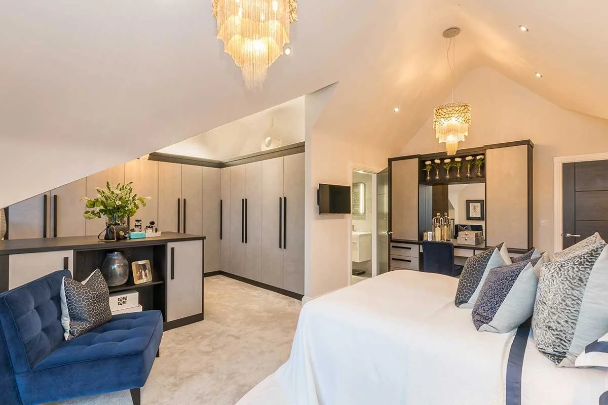 A modern bedroom with white walls, a sloped ceiling, and a large bed with multiple patterned pillows. There is a blue sofa with pillows, a dark wood desk with bottles and plants, and built-in cabinets with tall black handles. The room has two hanging chandeliers, recessed ceiling lights, and a small flat-screen TV.