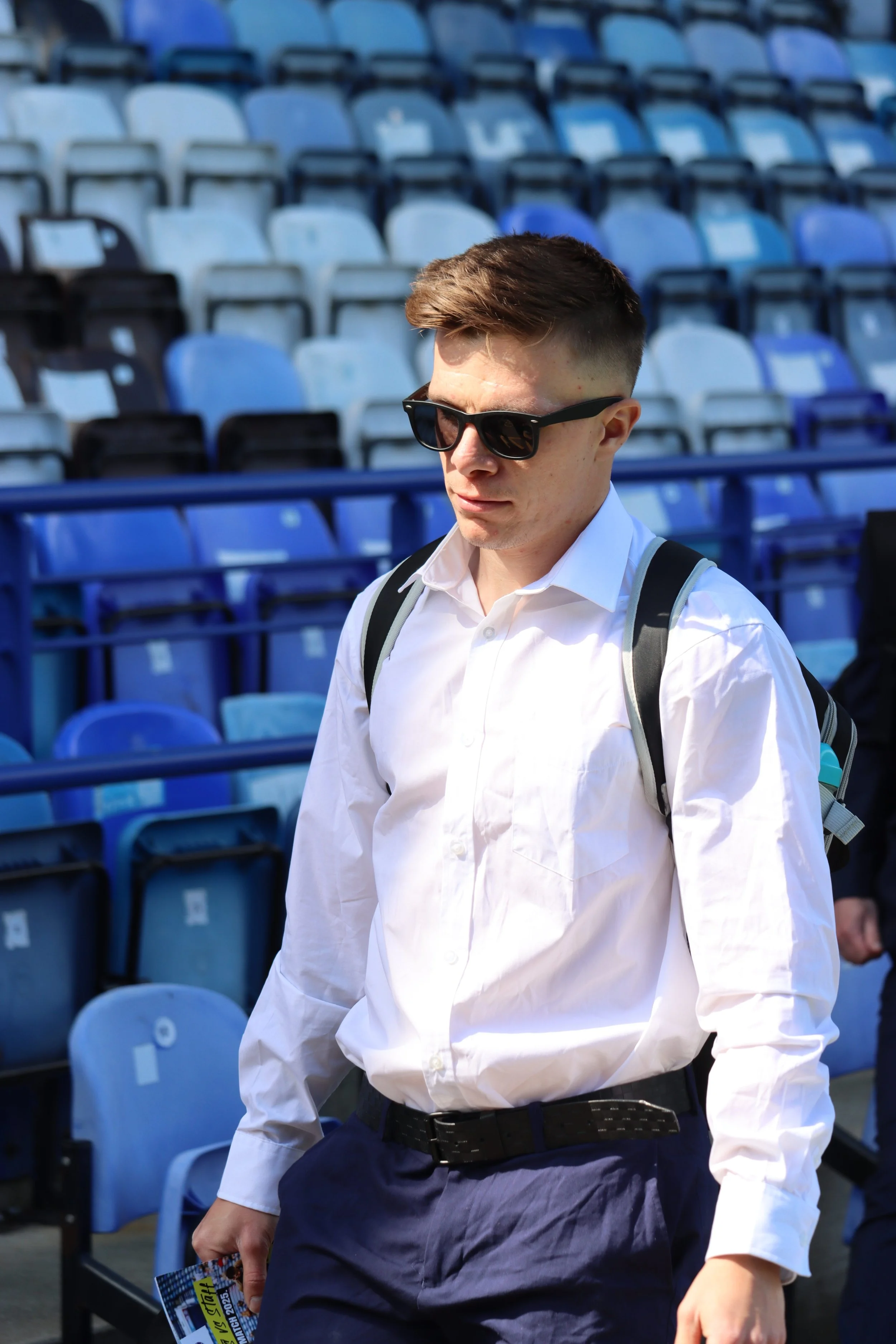 A young man with short brown hair, wearing black sunglasses, a white button-up shirt, and dark pants, walking in a stadium with empty blue and black seats.