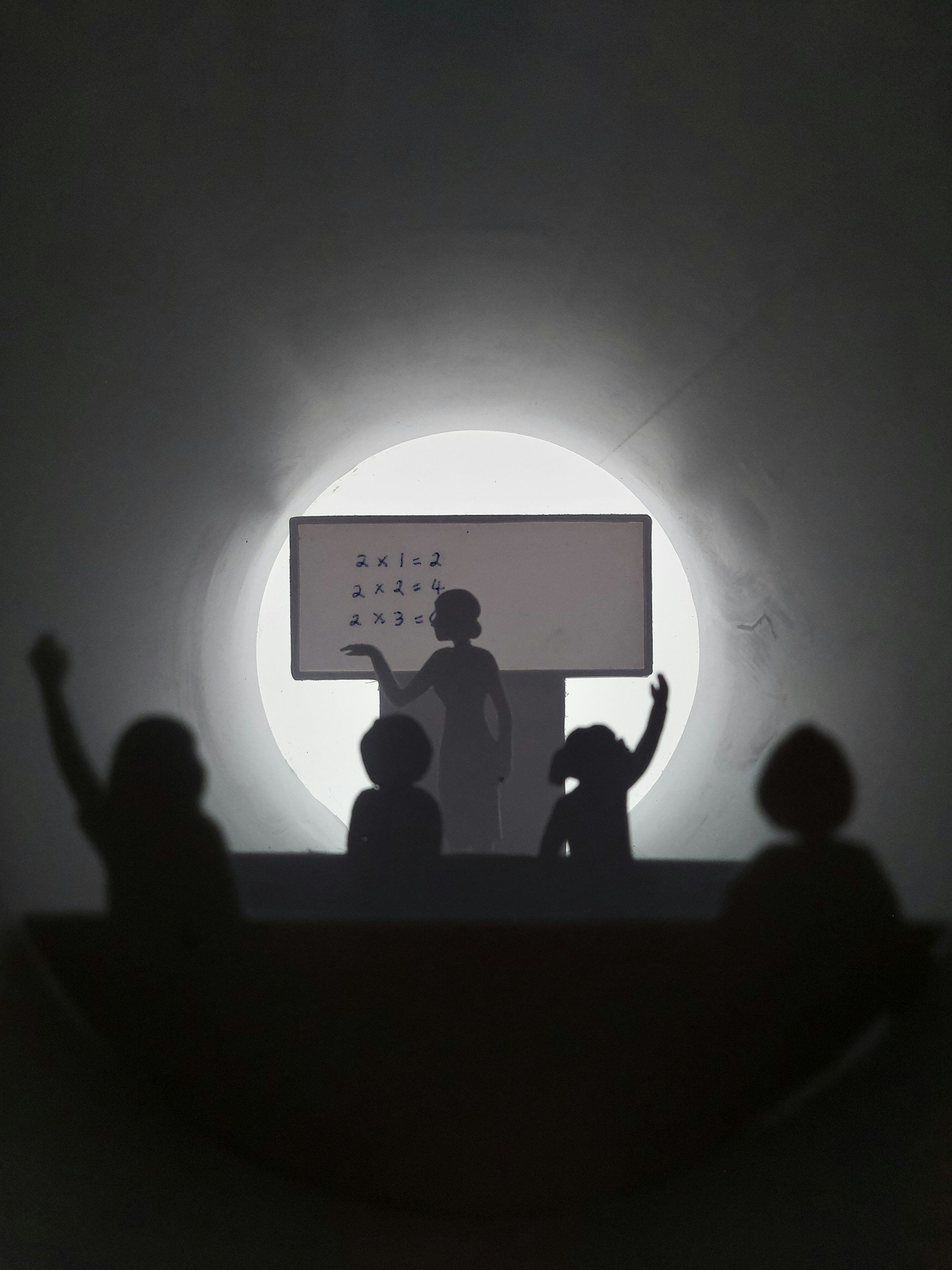 Silhouettes of children raising hands in front of a teacher at a blackboard, with the blackboard silhouette in front of a bright circular background.