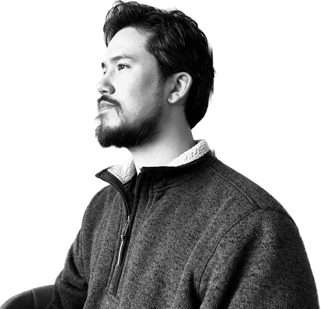 A black-and-white portrait of a man with a beard and mustache, looking sideways, wearing a zip-up sweater with a sherpa collar.