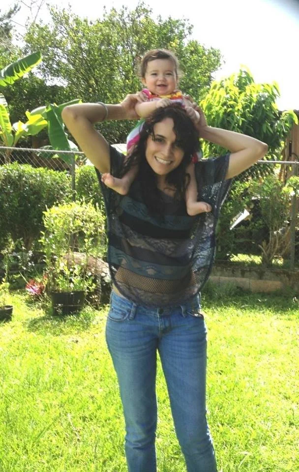 A woman with dark curly hair and a black shirt holding a smiling toddler girl with light brown hair on her shoulders outdoors in a grassy backyard with trees and bushes.
