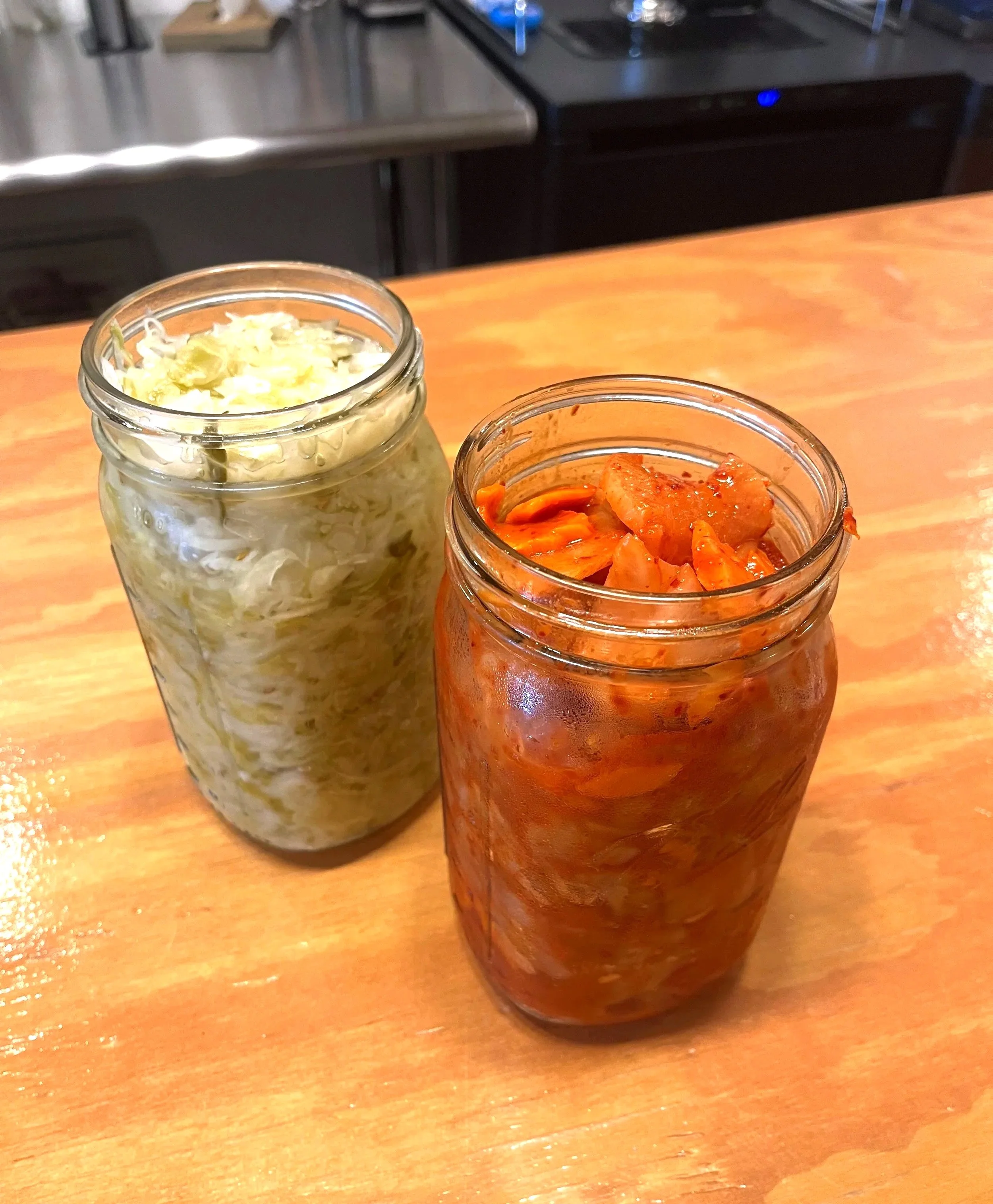 Two glass jars of kimchi placed on a wooden table, one with cabbage and the other with spicy fermented vegetables.