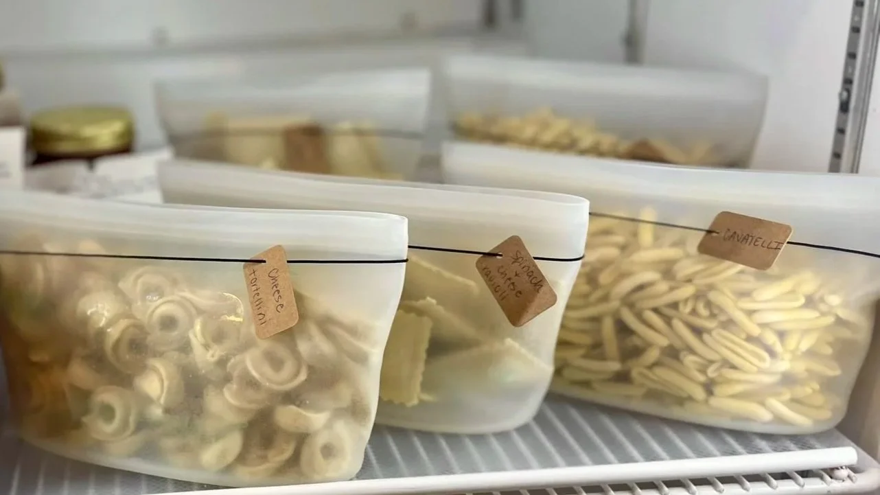 Clear reusable bags filled with dried pasta varieties labeled with handwritten tags inside a fridge or freezer.