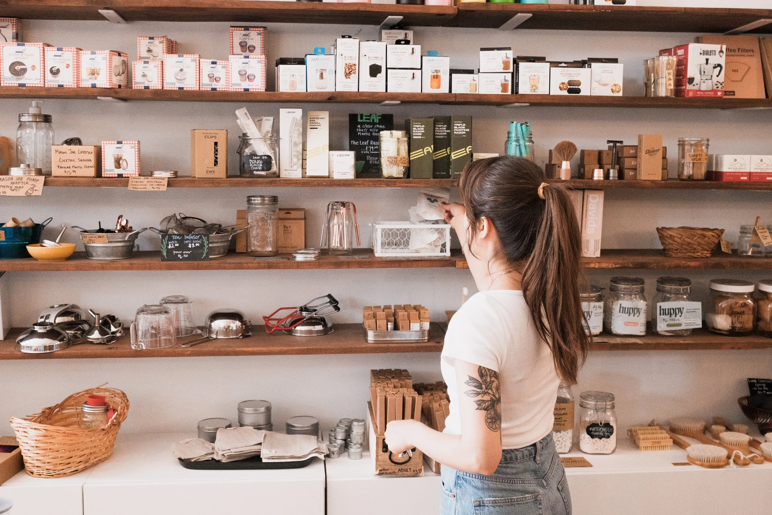 A woman with a tattoo on her arm and tied-back hair shopping at a store with wooden shelves filled with various boxes, jars, and bottles of products.