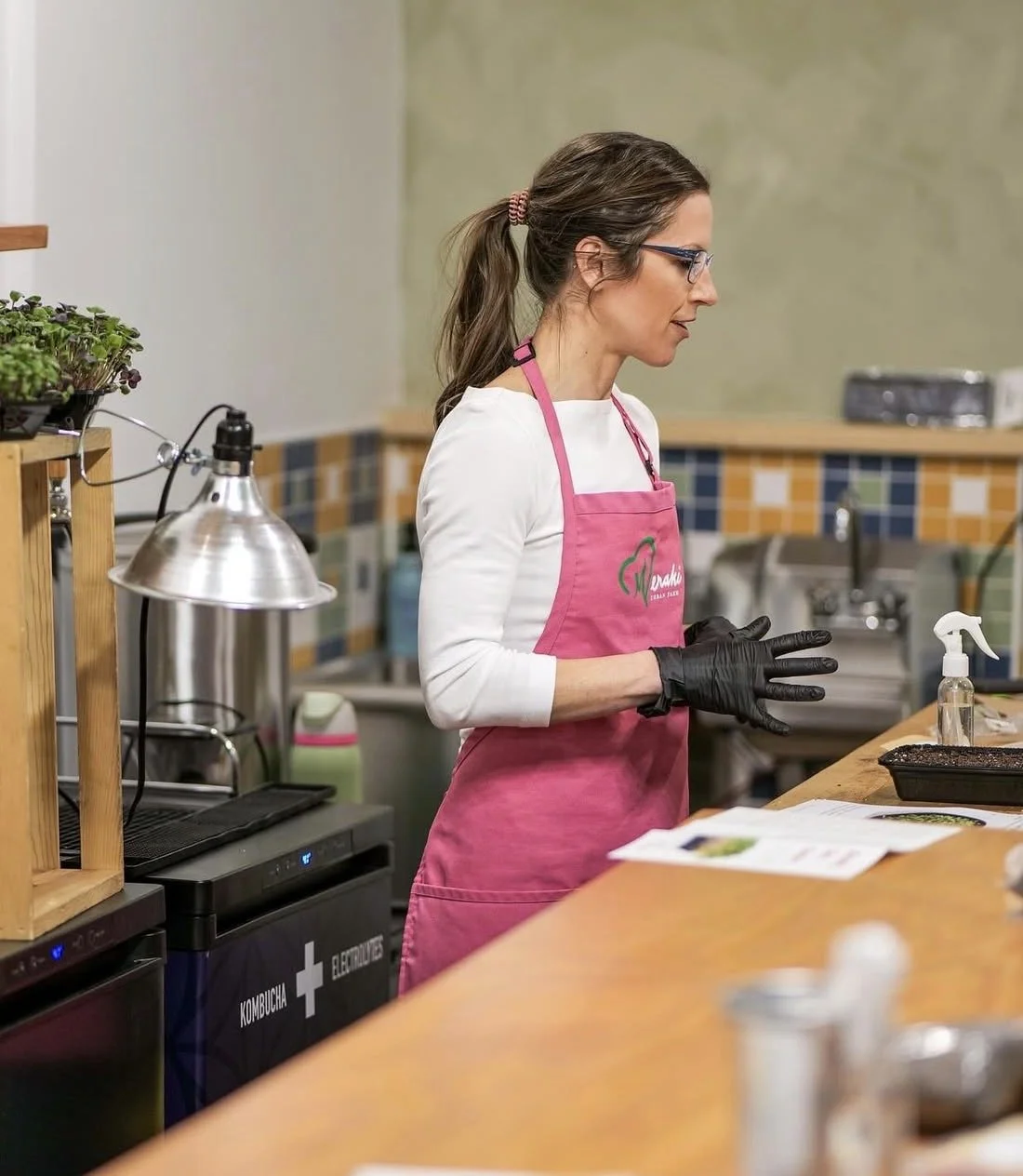 A woman wearing glasses, a white long sleeve shirt, and a pink apron, standing in a kitchen, preparing food.