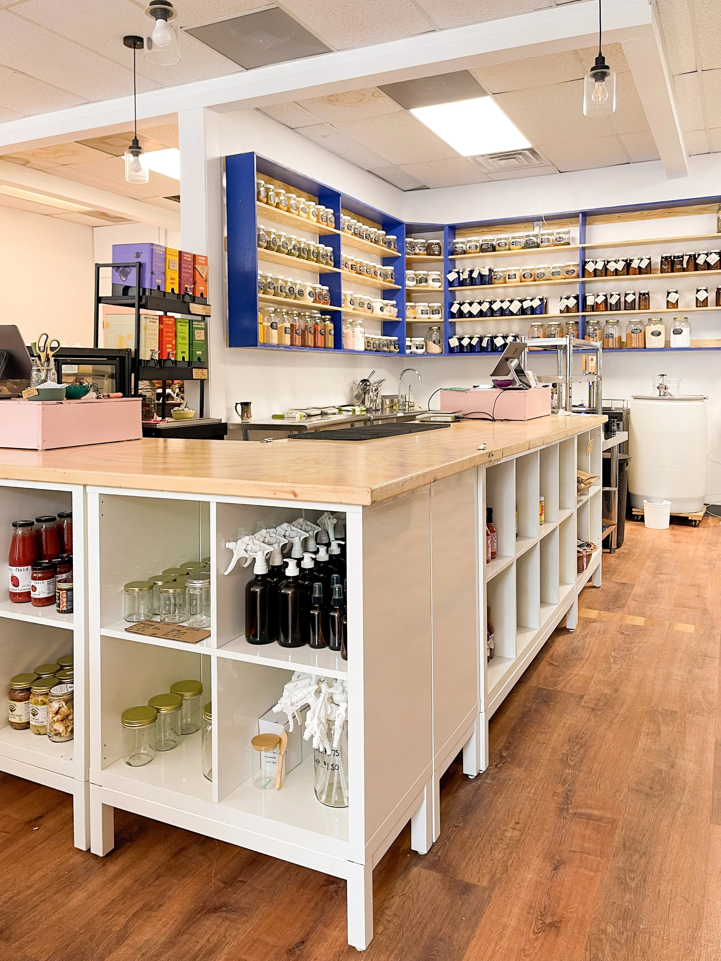 A display counter in a store with shelves of jars and bottles containing various spices, herbs, and ingredients, with a cash register, some office supplies.