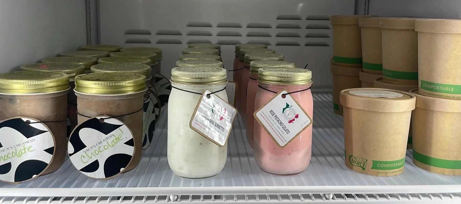 Jars of ice cream in a refrigerator, including flavors like chocolate, basil sorbet, and spinach gelato, with eco-friendly, compostable containers stacked on the right side.
