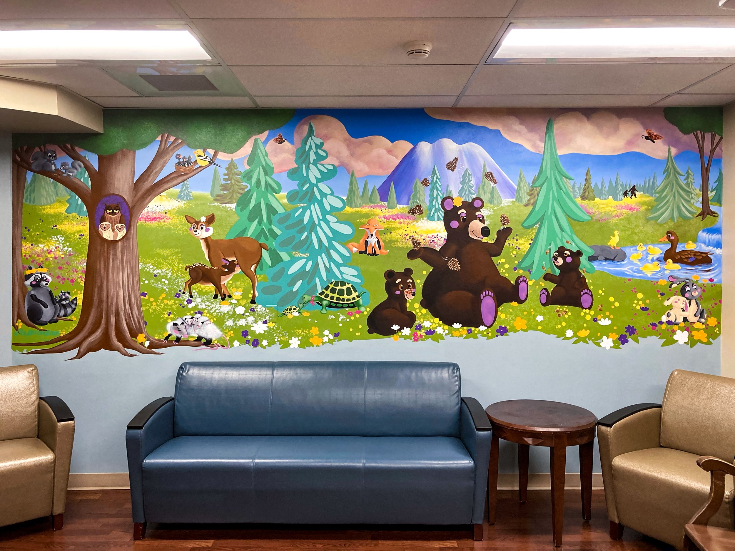 Colorful mural depicting a forest scene with cartoon animals like bears, deer, raccoons, and ducks. The mural includes trees, flowers, mountains, and a stream, providing a cheerful and whimsical nature setting. Located above a blue couch between two