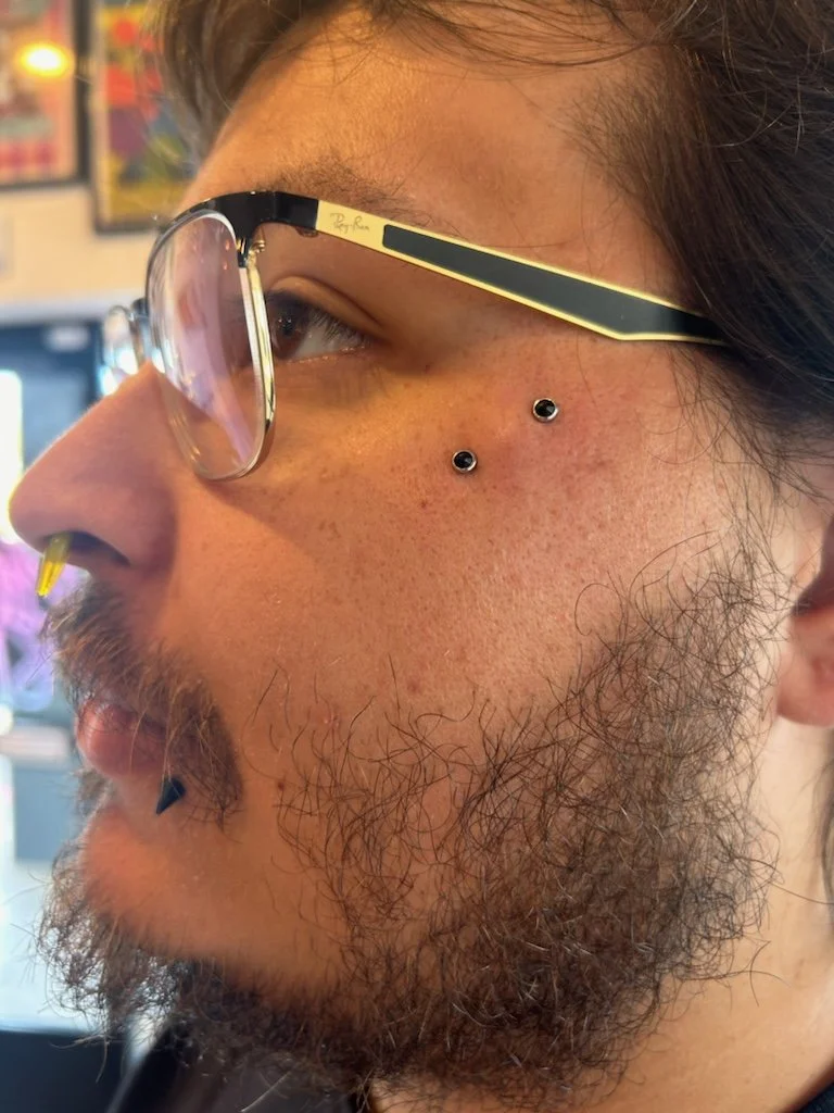 Close-up of a man's face showing glasses, facial piercings, and facial hair, with a blurred background.
