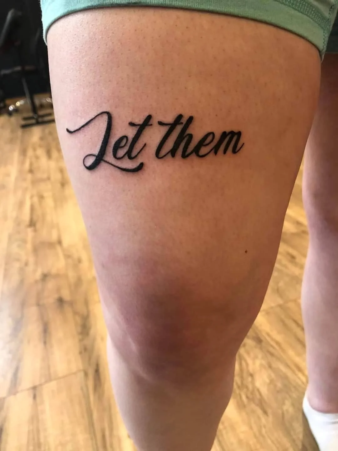 A tattoo on a person's thigh that says 'Let Them' in black cursive font.