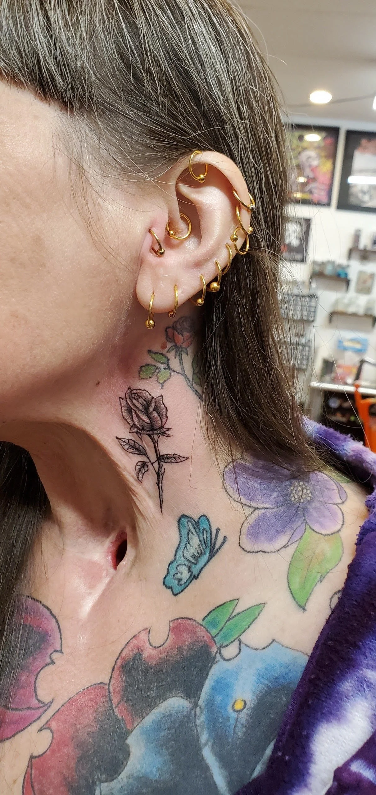 Close-up of a person's ear with multiple gold earrings and a neck tattoo of a rose, flowers, and a butterfly.