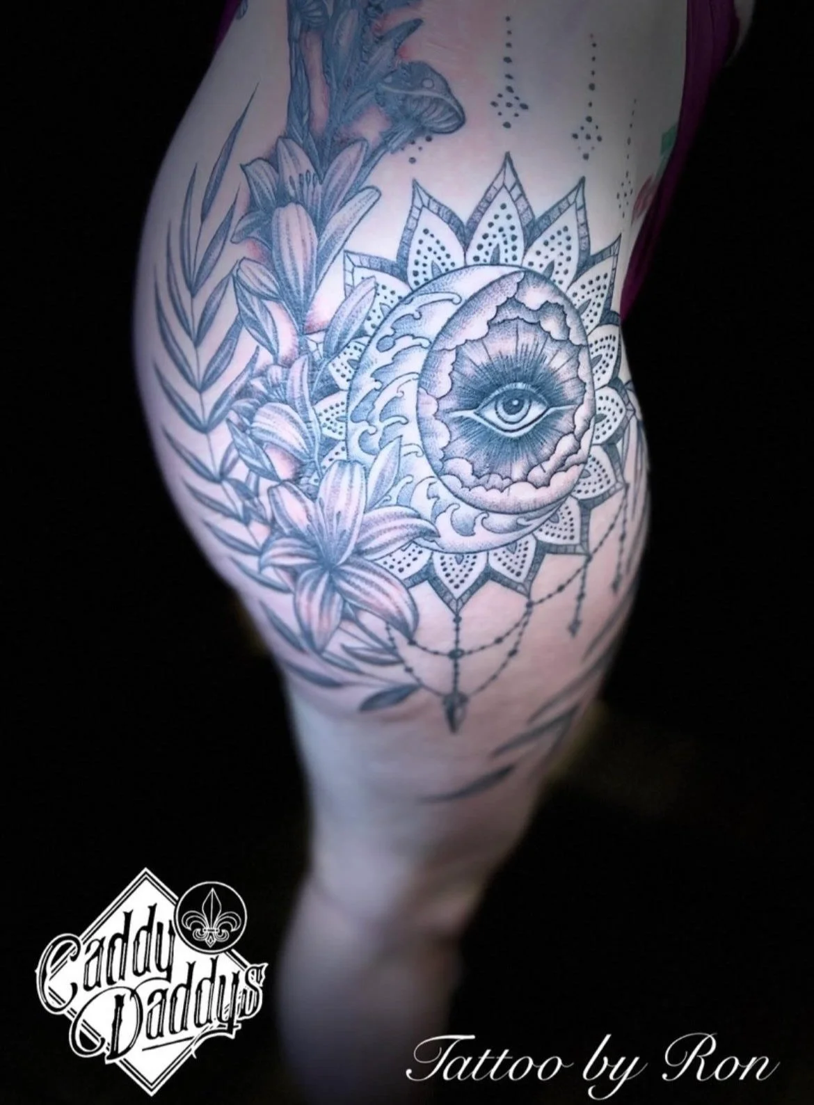 Tattoo of an eye within a moon surrounded by flowers and a mandala, on a person's thigh.
